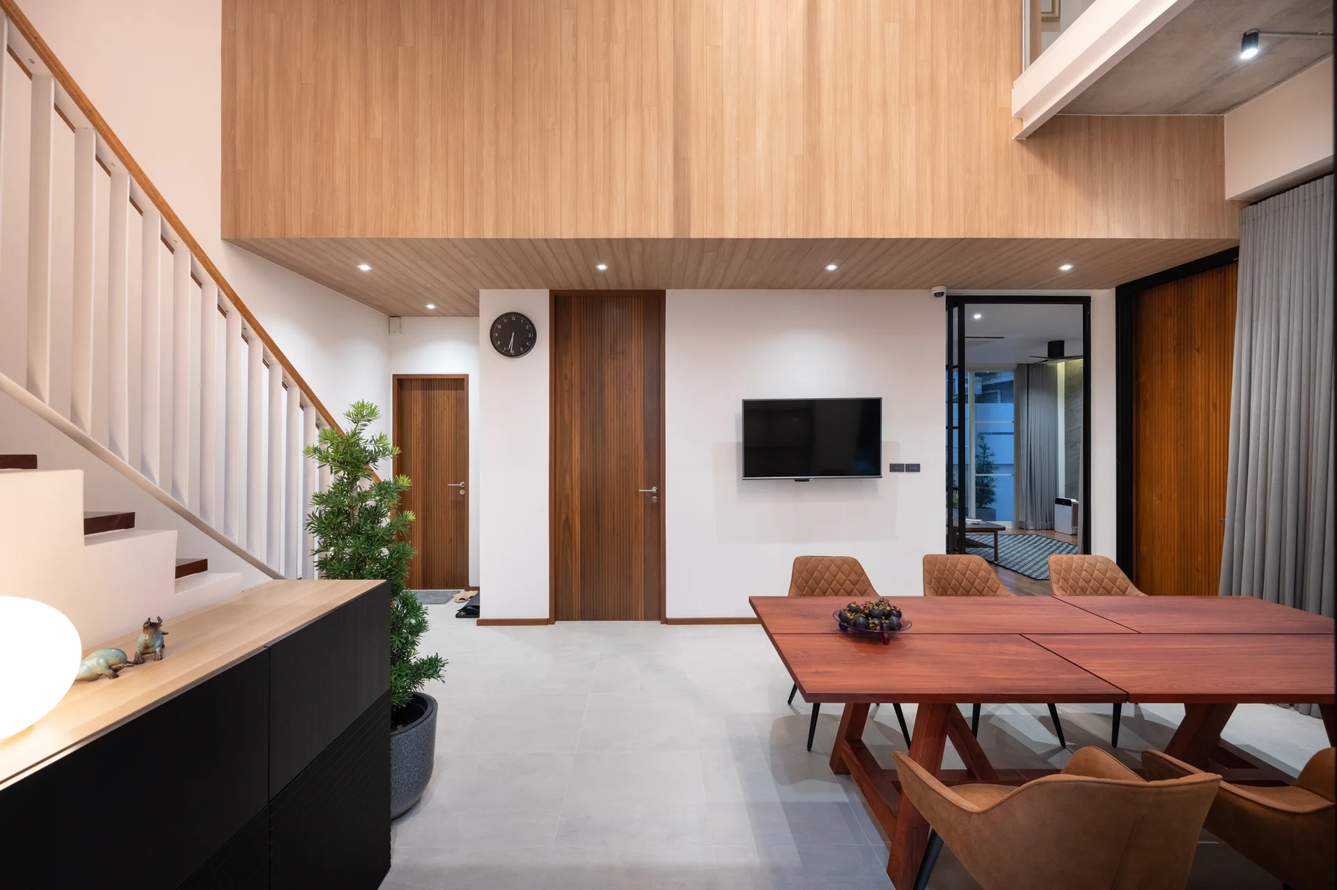 A modern dining room featuring a large reddish-brown wood table, tan upholstered chairs, white walls, and a warm wood-paneled upper wall near a staircase.