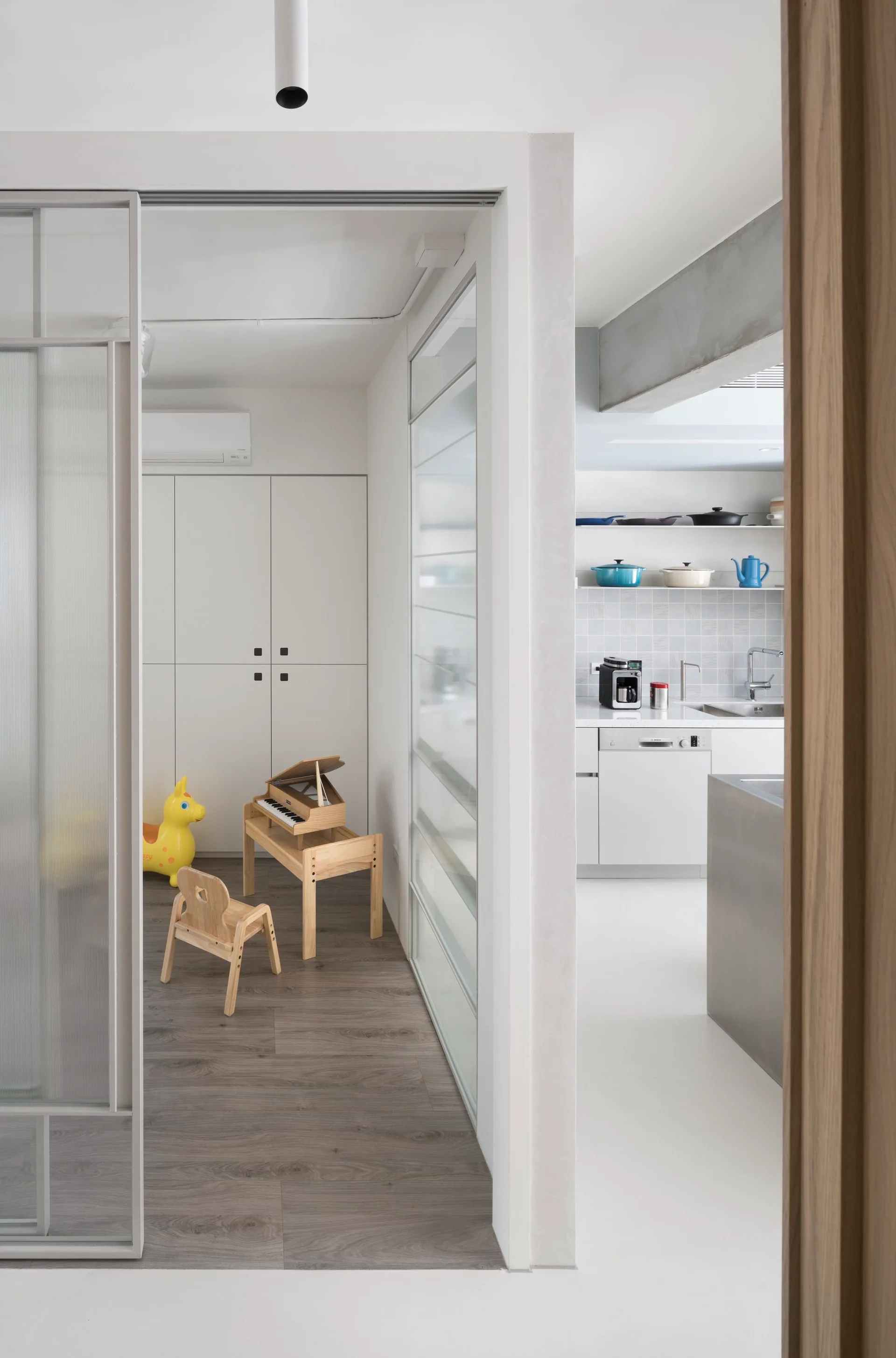 A view through a sliding glass partition of a playroom with a toy piano next to a minimalist white kitchen with open shelving.