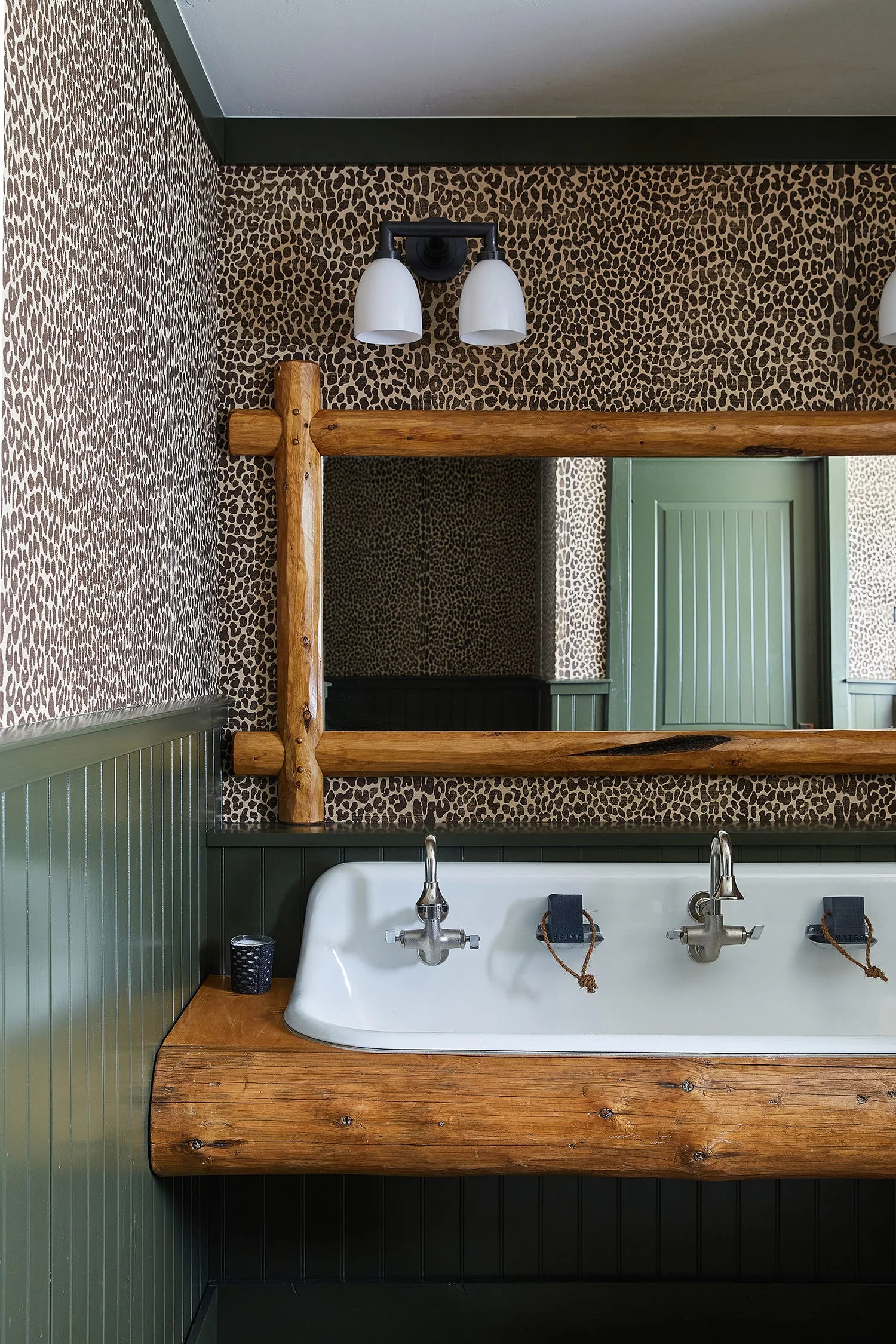 A white ceramic trough sink rests on a log base, paired with green beadboard wainscoting, leopard print wallpaper, and a log-framed mirror.
