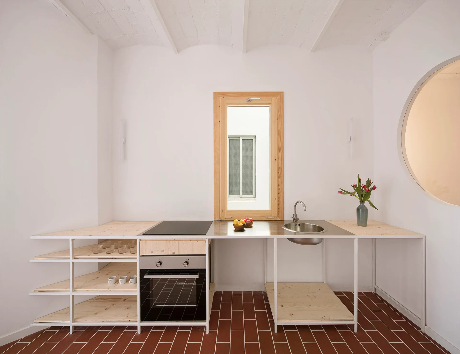 A minimalist kitchen featuring an open-frame plywood console with a stainless steel sink, integrated cooktop, and terracotta brick tile floor.