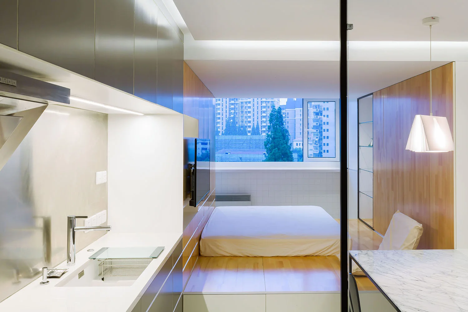 A modern studio apartment interior featuring a white-covered bed on a raised wooden platform next to a grey and white kitchenette.