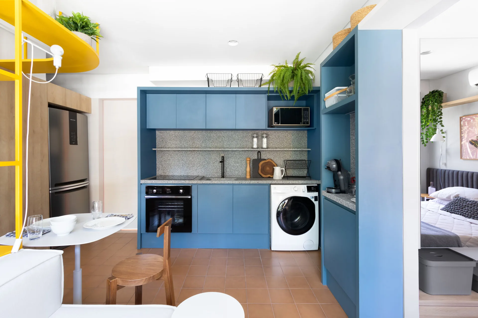 A compact kitchen features matte blue cabinetry, a speckled gray backsplash, a front-loading washing machine, and terracotta-colored floor tiles.