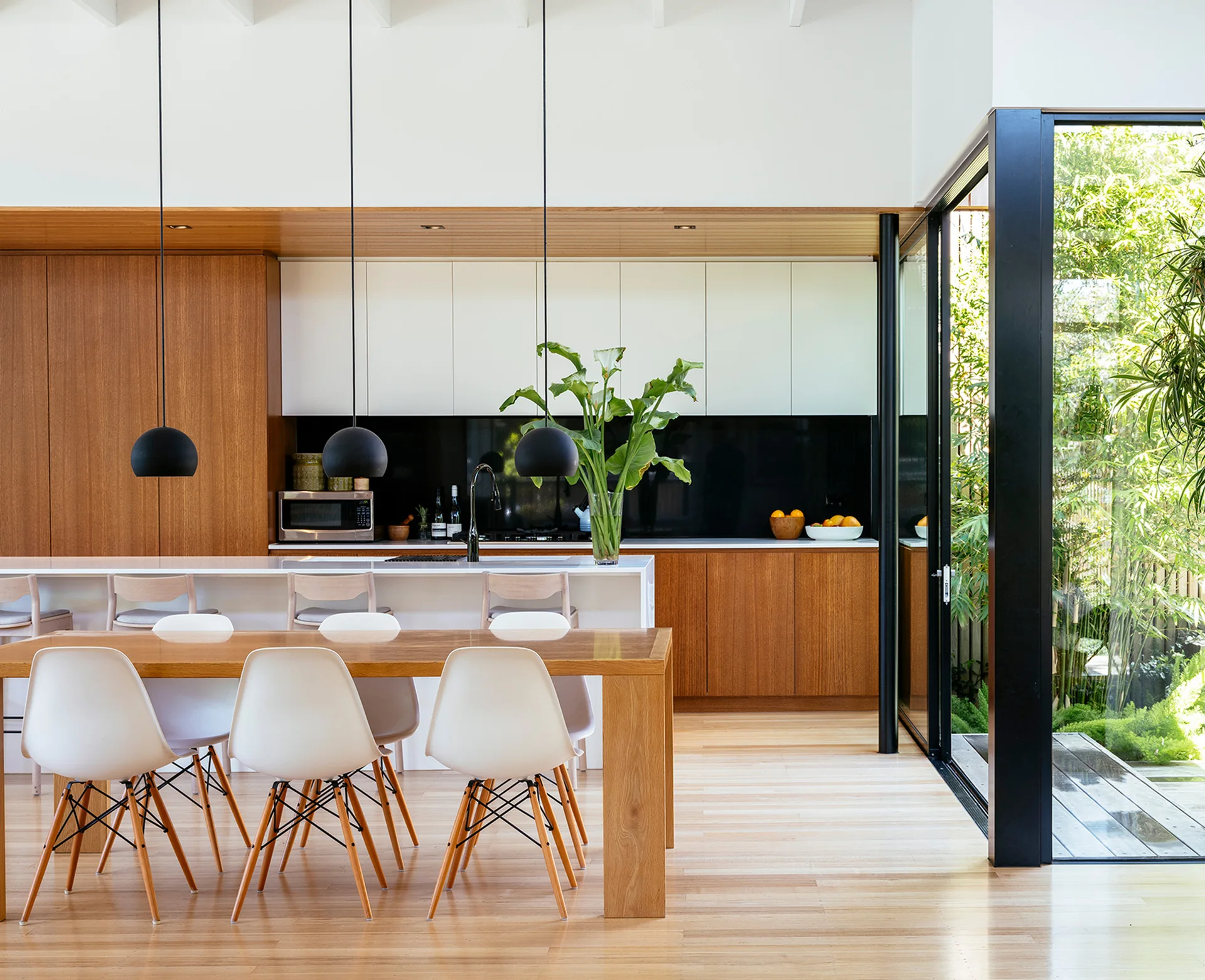 A modern open-concept kitchen and dining area features warm wood cabinetry, a white island, black pendant lights, and a sliding glass door opening to a garden.