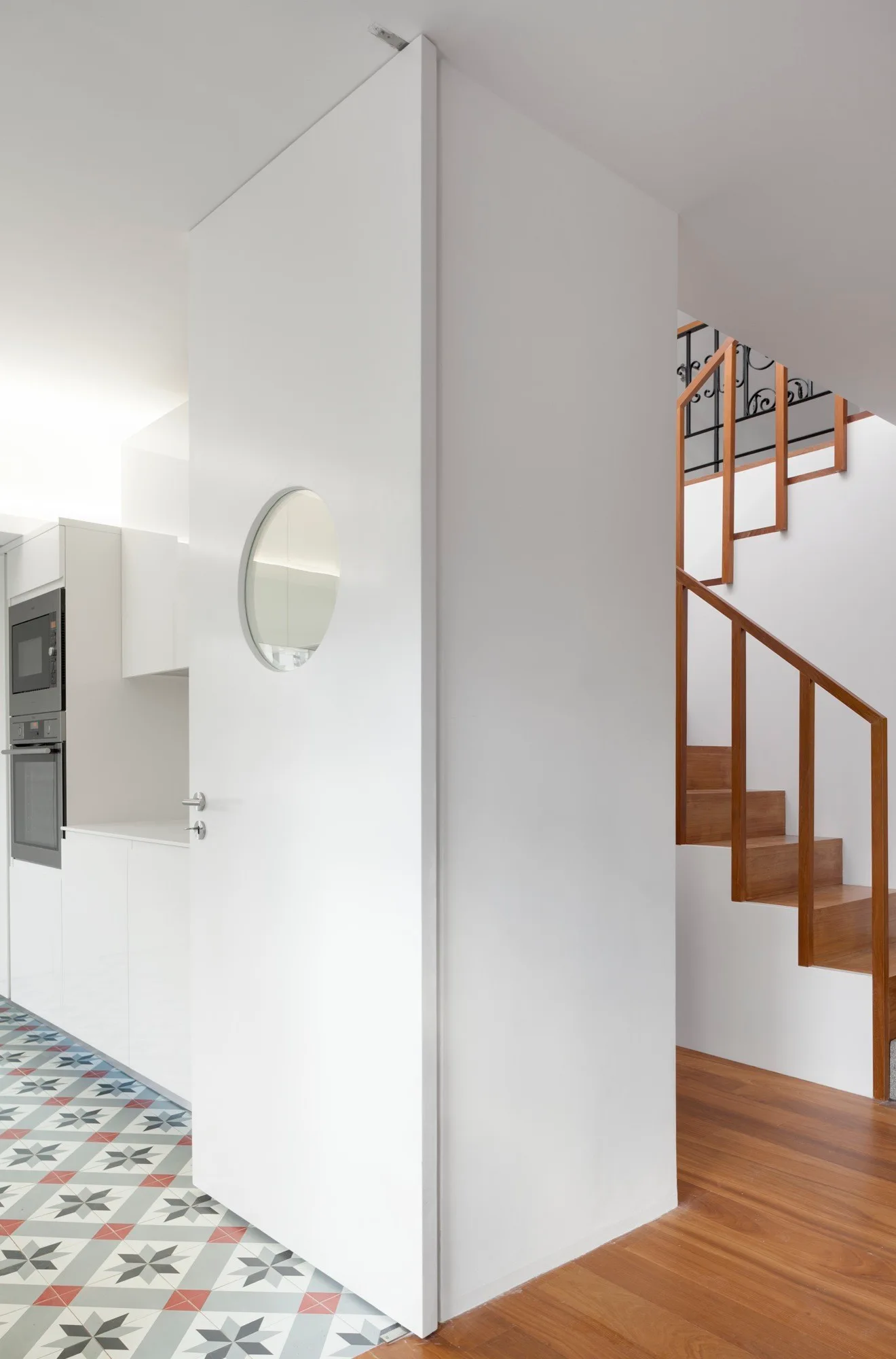 A white minimalist door with a circular window stands between a kitchen with geometric floor tiles and a wooden staircase.