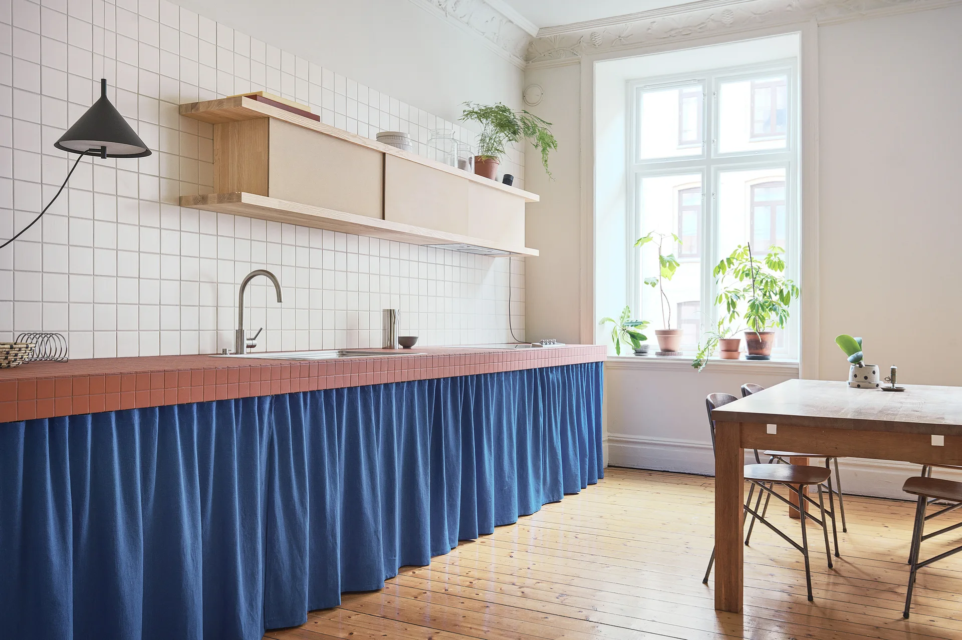 A kitchen features a long terracotta tiled counter with a blue fabric skirt, white square wall tiles, and a natural wood dining table.