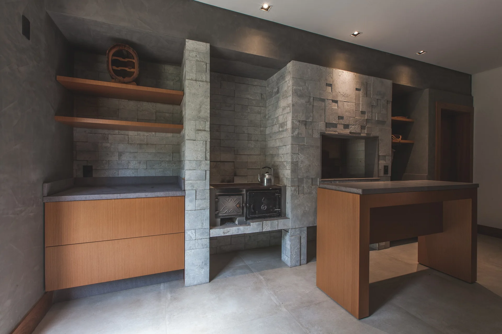 Modern kitchen with a central grey stone block fireplace, flat-front wood cabinetry, floating shelves, and a matching wood prep island.