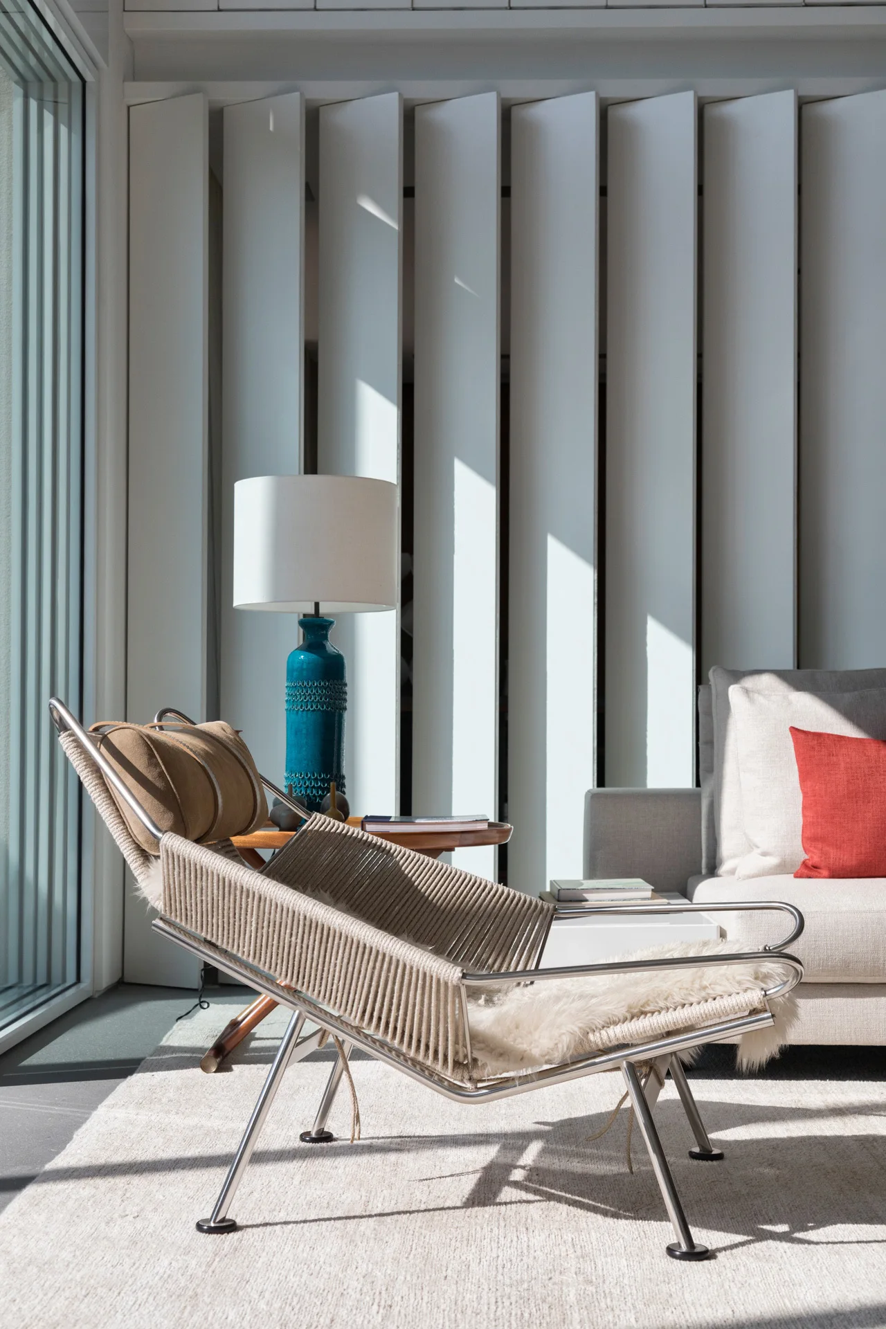 A modern corded lounge chair with a sheepskin throw sits in a sunlit room featuring vertical white louvered panels and a blue ceramic lamp.