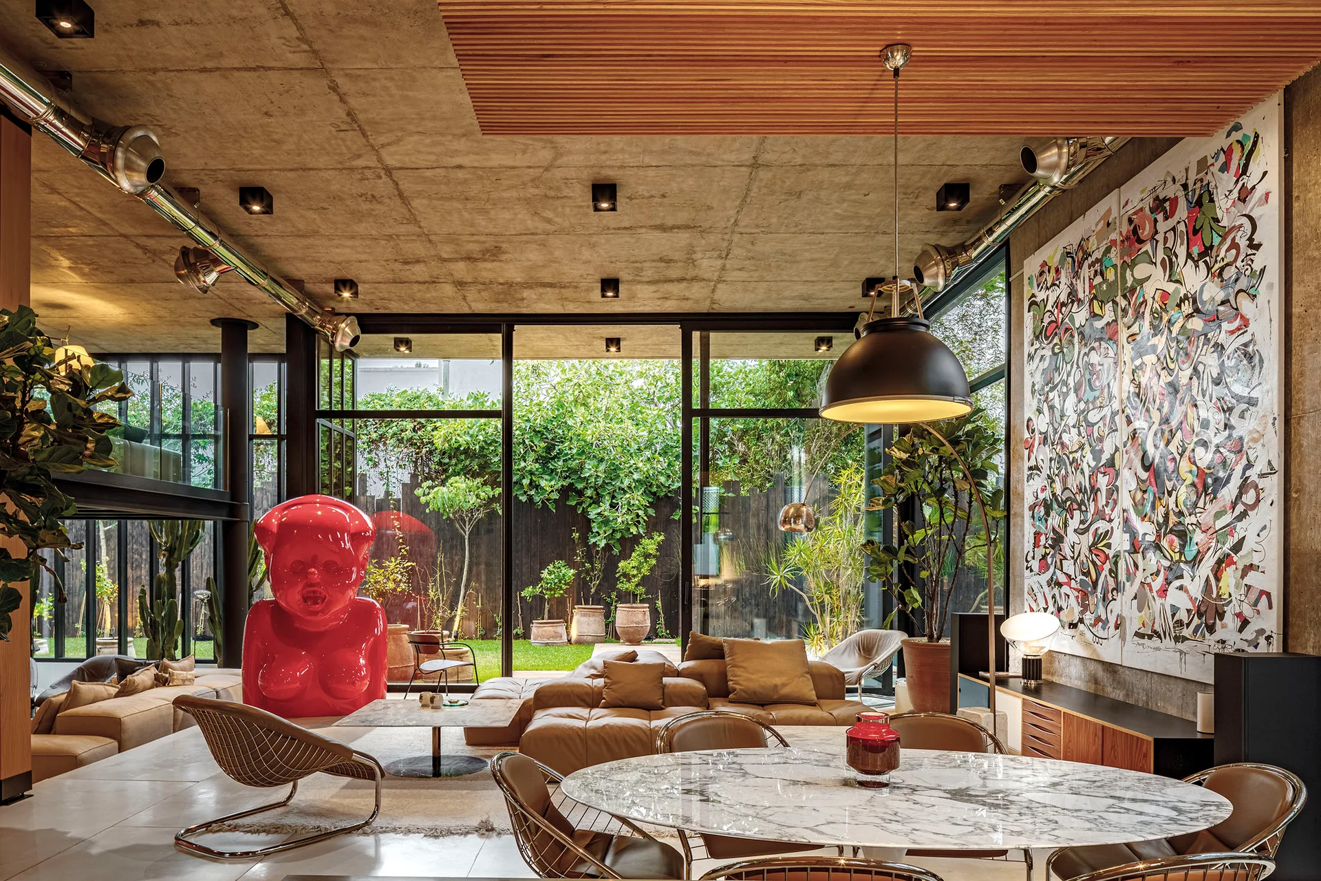 An industrial living room with an exposed concrete ceiling, floor-to-ceiling windows showing a garden, a tan leather sofa, and a large red sculpture.