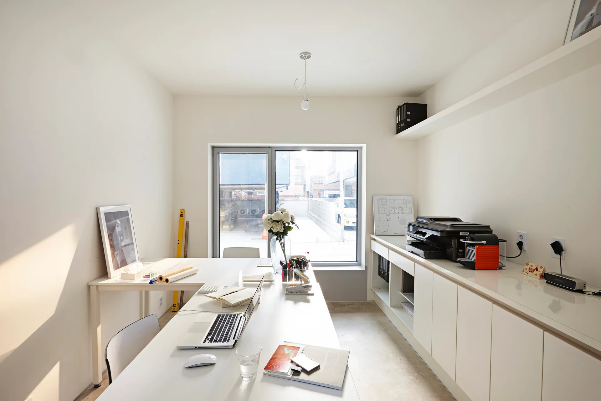A minimalist home office features a long white desk with a laptop, matte white built-in cabinets, a floating shelf, and a large window.