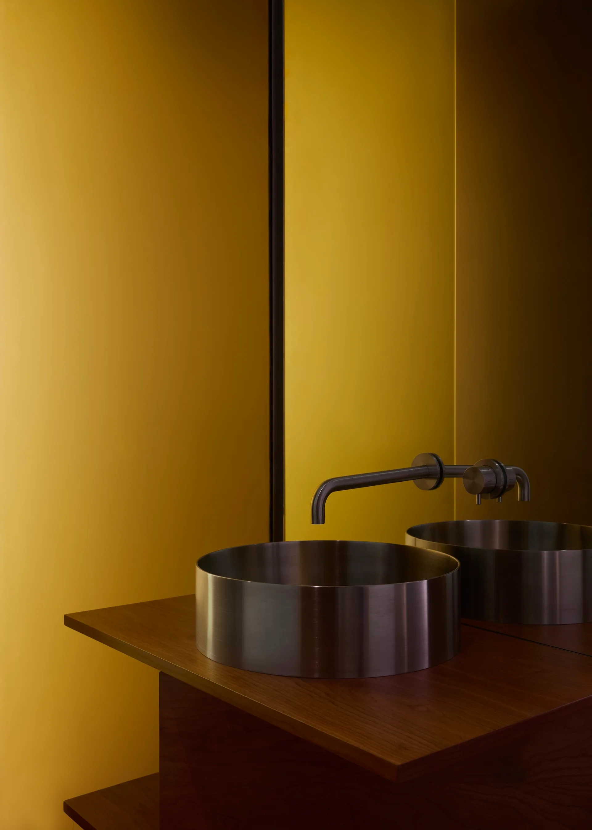 A circular brushed metal vessel sink with a wall-mounted faucet on a wood vanity in front of a bright yellow wall and mirror.