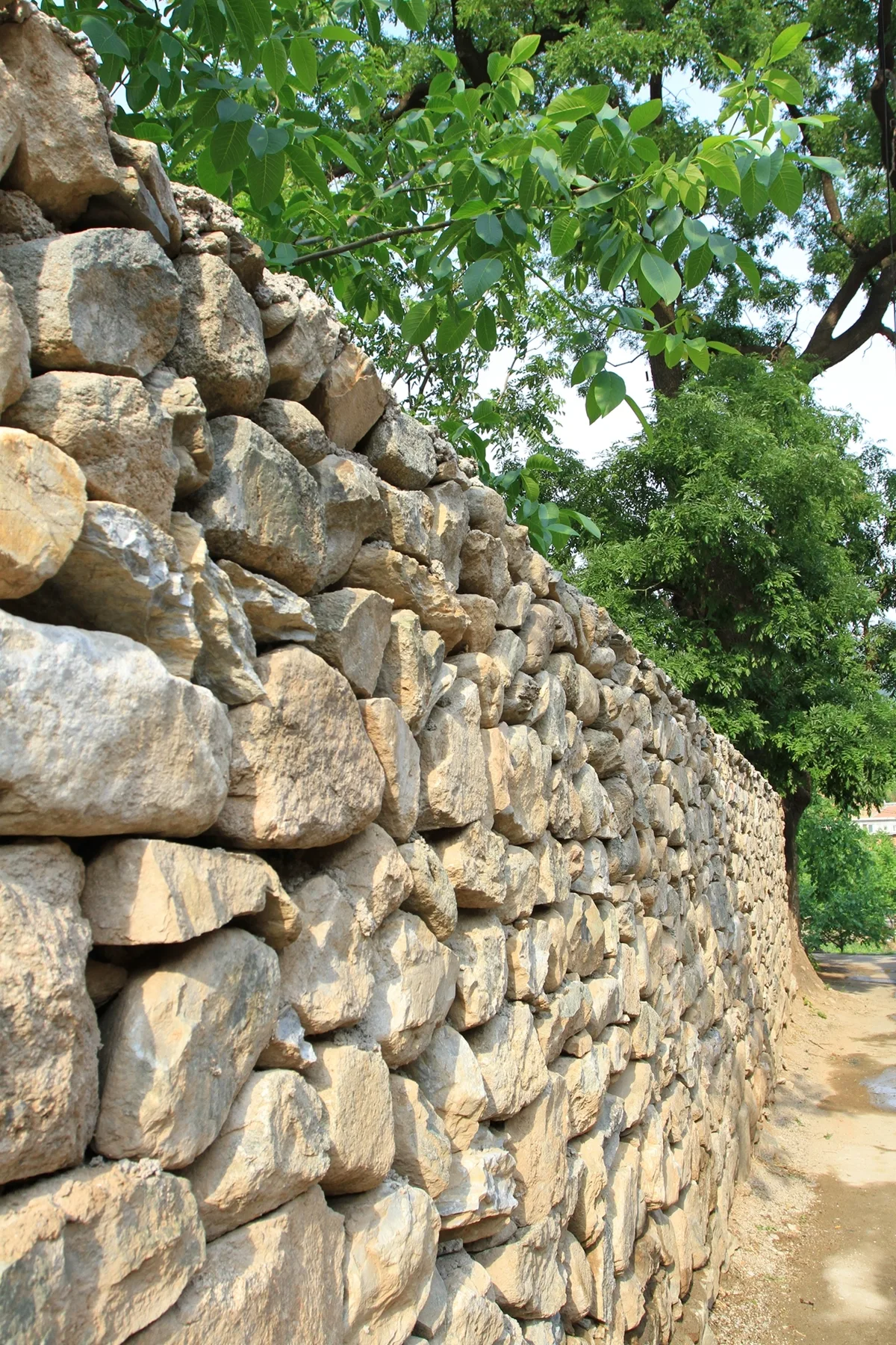 A rustic stacked stone retaining wall constructed from irregular beige and grey rocks runs alongside a dirt path under green trees.