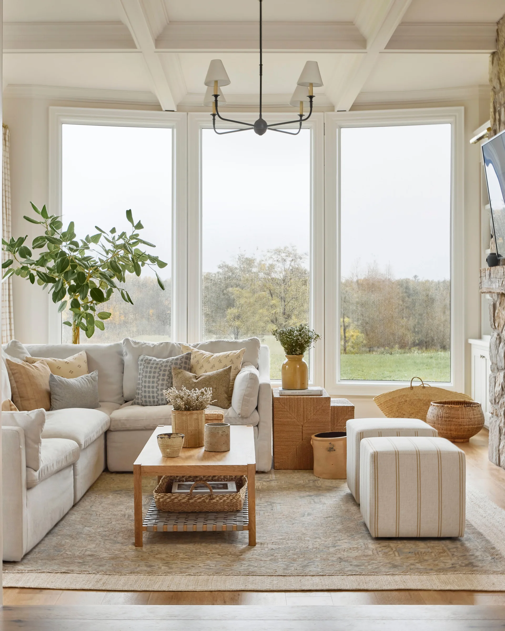 A bright living room features a cream sectional sofa, a wood coffee table with striped ottomans, and large windows looking out to trees.