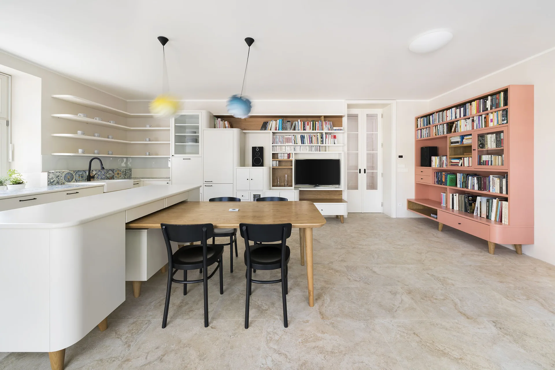An open-concept kitchen features a white island with an attached wooden dining table, black chairs, and a large pink modular bookshelf on stone floor tiles.