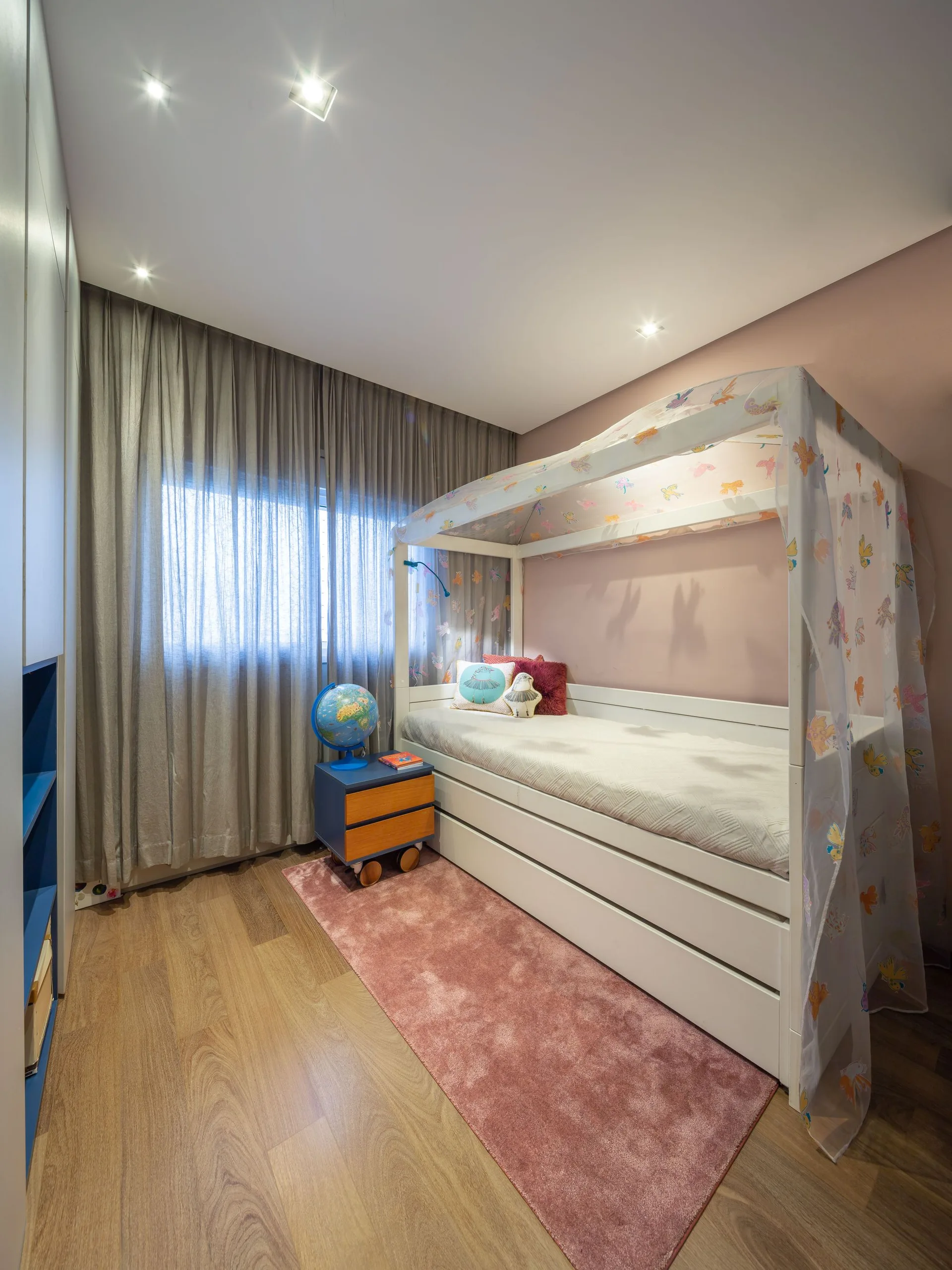 A children's bedroom features a white canopy bed with patterned netting against a pink wall, a matching pink runner rug, and grey sheer curtains.