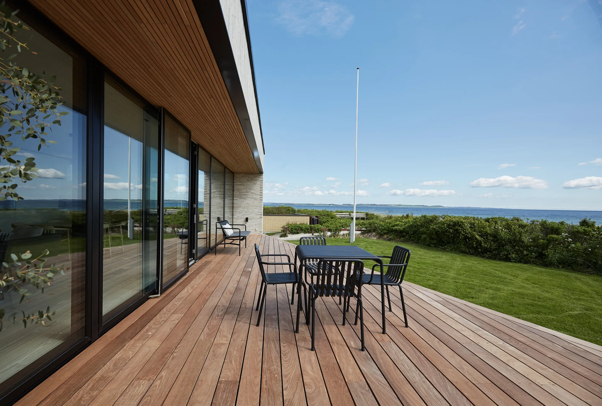 A modern wooden deck featuring black metal patio furniture and large glass sliding doors overlooking a grassy lawn and the sea.