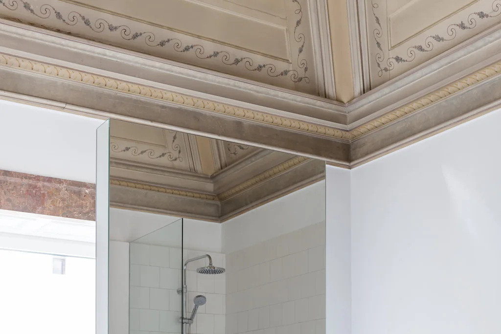 An ornate painted plaster ceiling with scroll patterns sits above a large mirror reflecting a white subway tile shower and chrome fixtures.