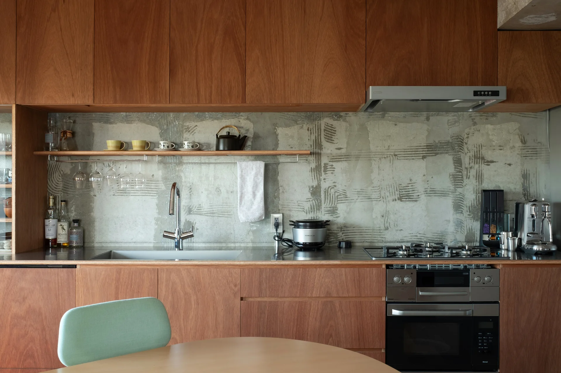 A modern kitchen featuring flat-front wood cabinetry, a raw concrete backsplash, a stainless steel countertop with sink, and a floating wooden shelf.