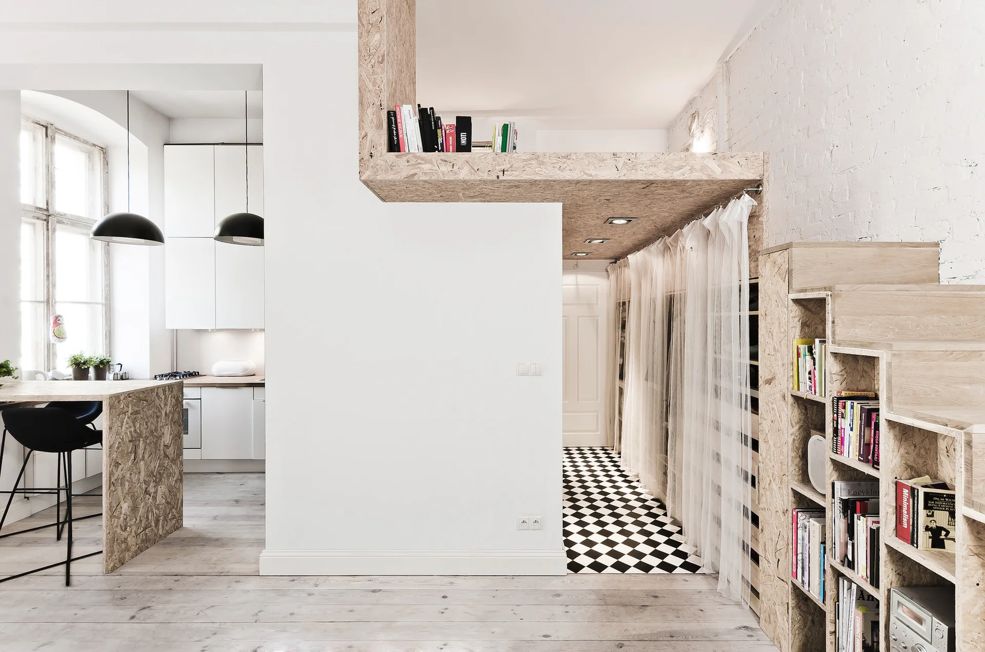 A modern white kitchen and hallway featuring a raw wood particle board loft structure, matching stepped bookshelf, and black-and-white checkered floor.