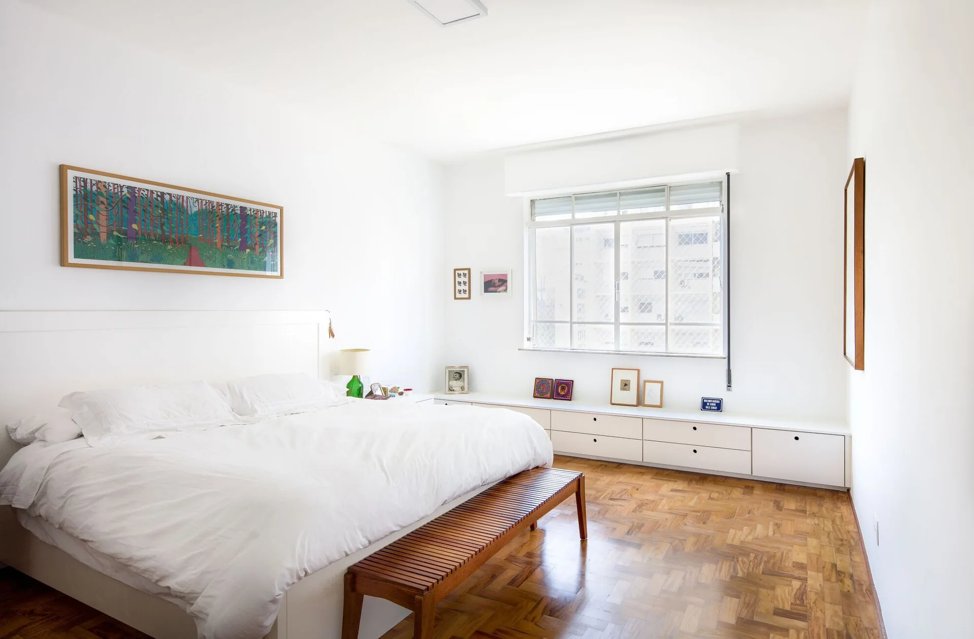 A bright white minimalist bedroom features a large bed, a slatted wooden bench, a parquet floor, and a long low-profile white dresser.