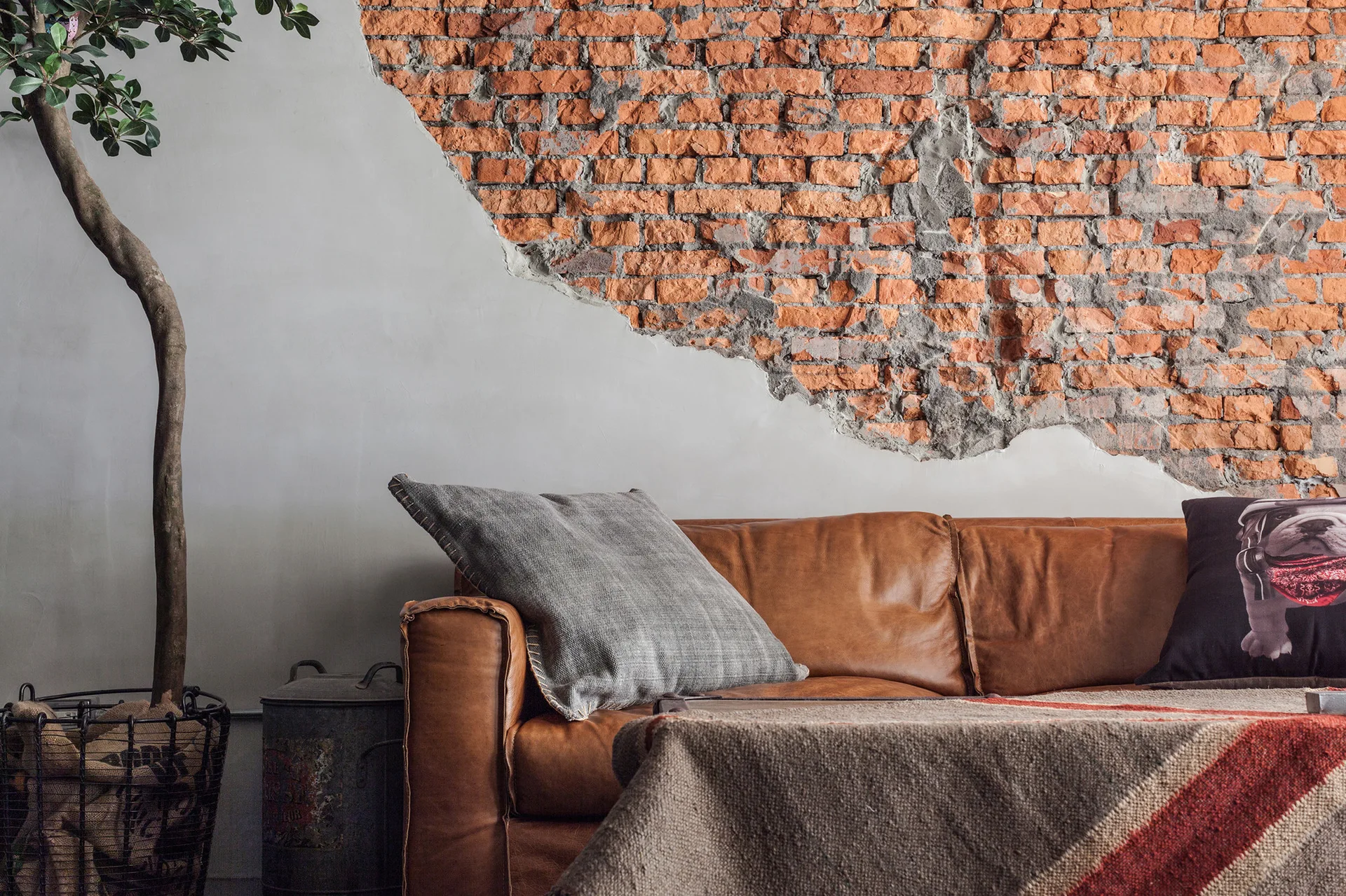 A brown leather sofa with textured pillows sits against a wall that transitions from smooth plaster to rough exposed red brick.