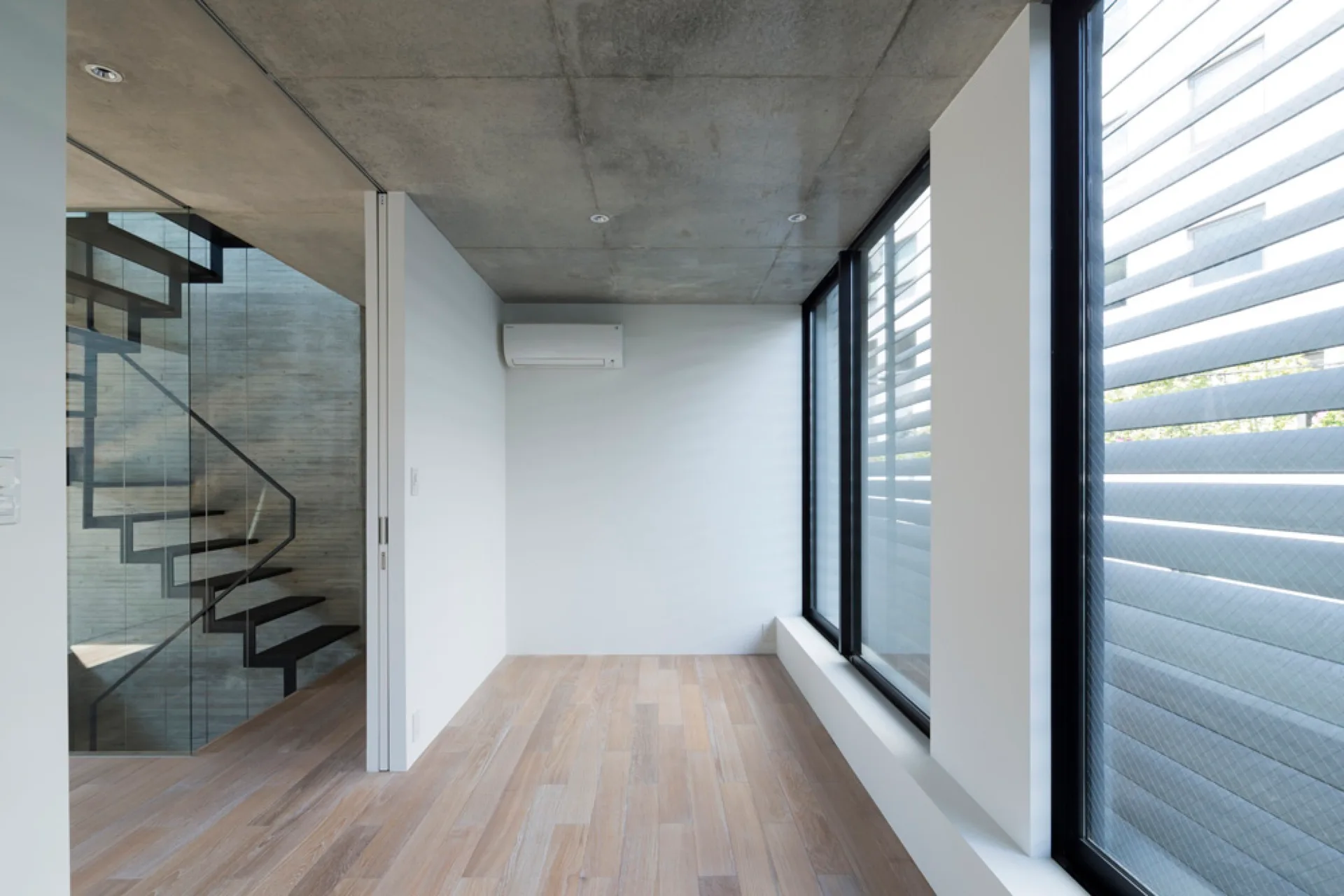 A minimalist room featuring an exposed concrete ceiling, light wood plank floors, large windows, and a black metal staircase enclosed in glass.