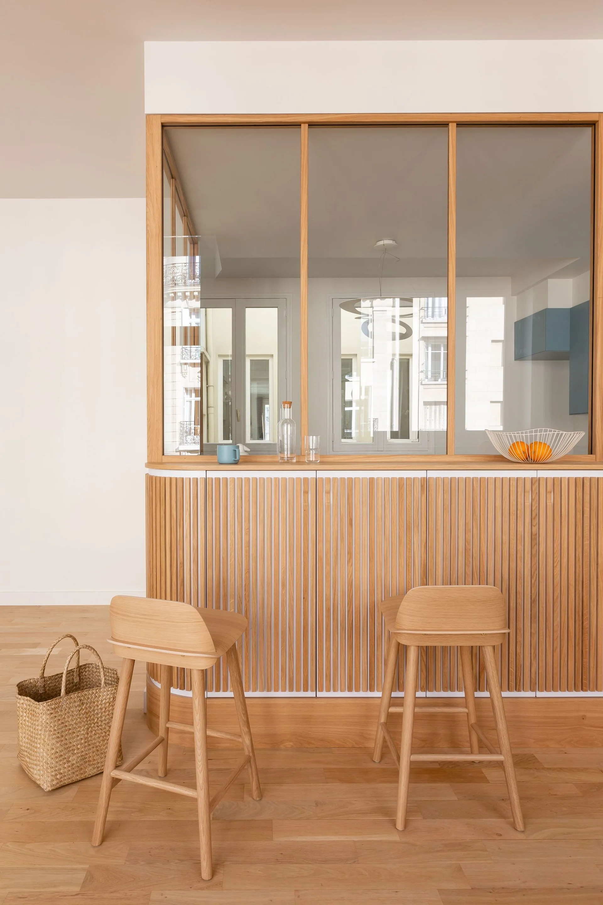 A light wood kitchen pass-through counter with vertical slatted paneling, a glass partition, and two matching wooden stools.