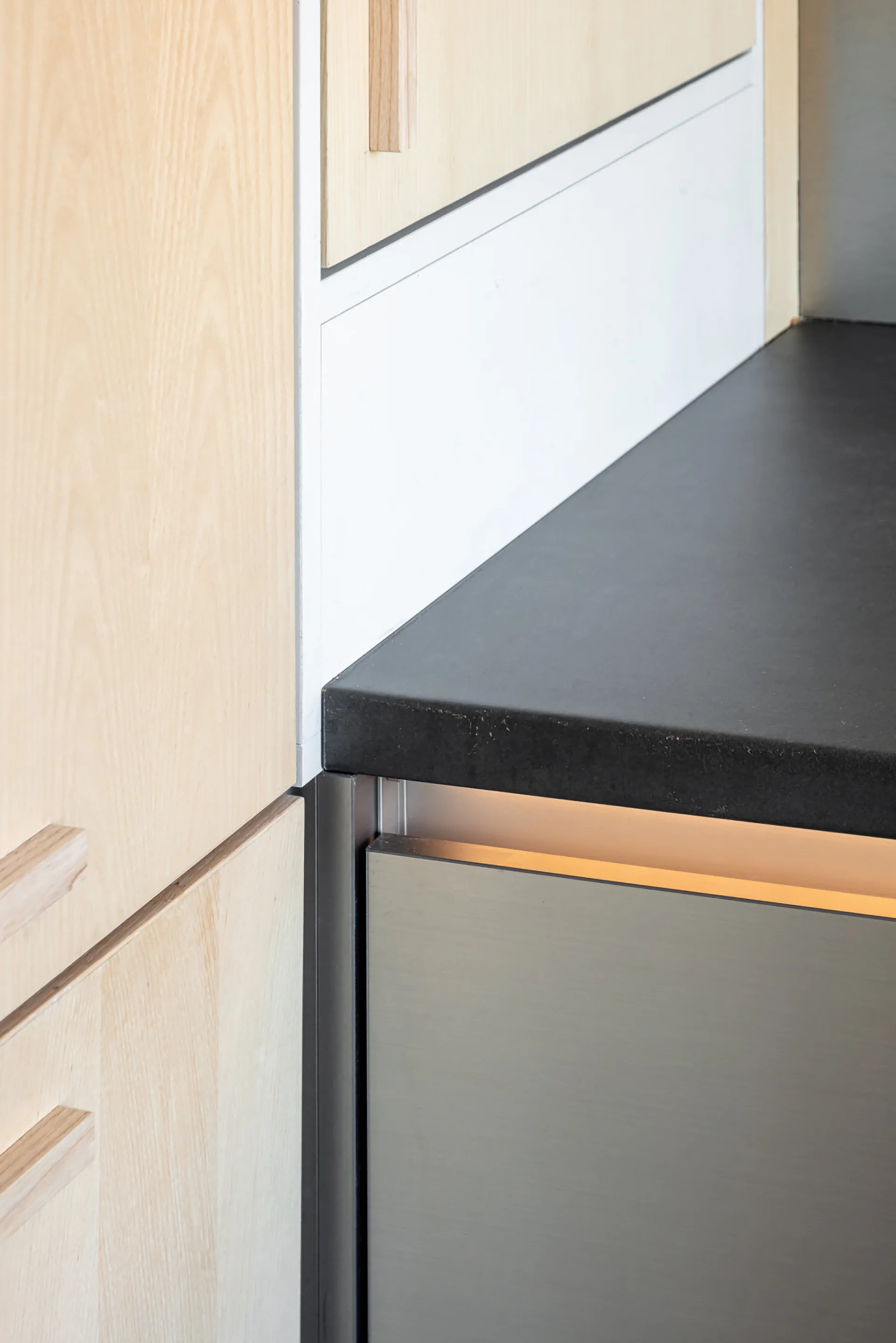 A close-up of light wood kitchen cabinetry alongside a dark stone countertop, a white backsplash, and a metallic appliance with under-cabinet lighting.