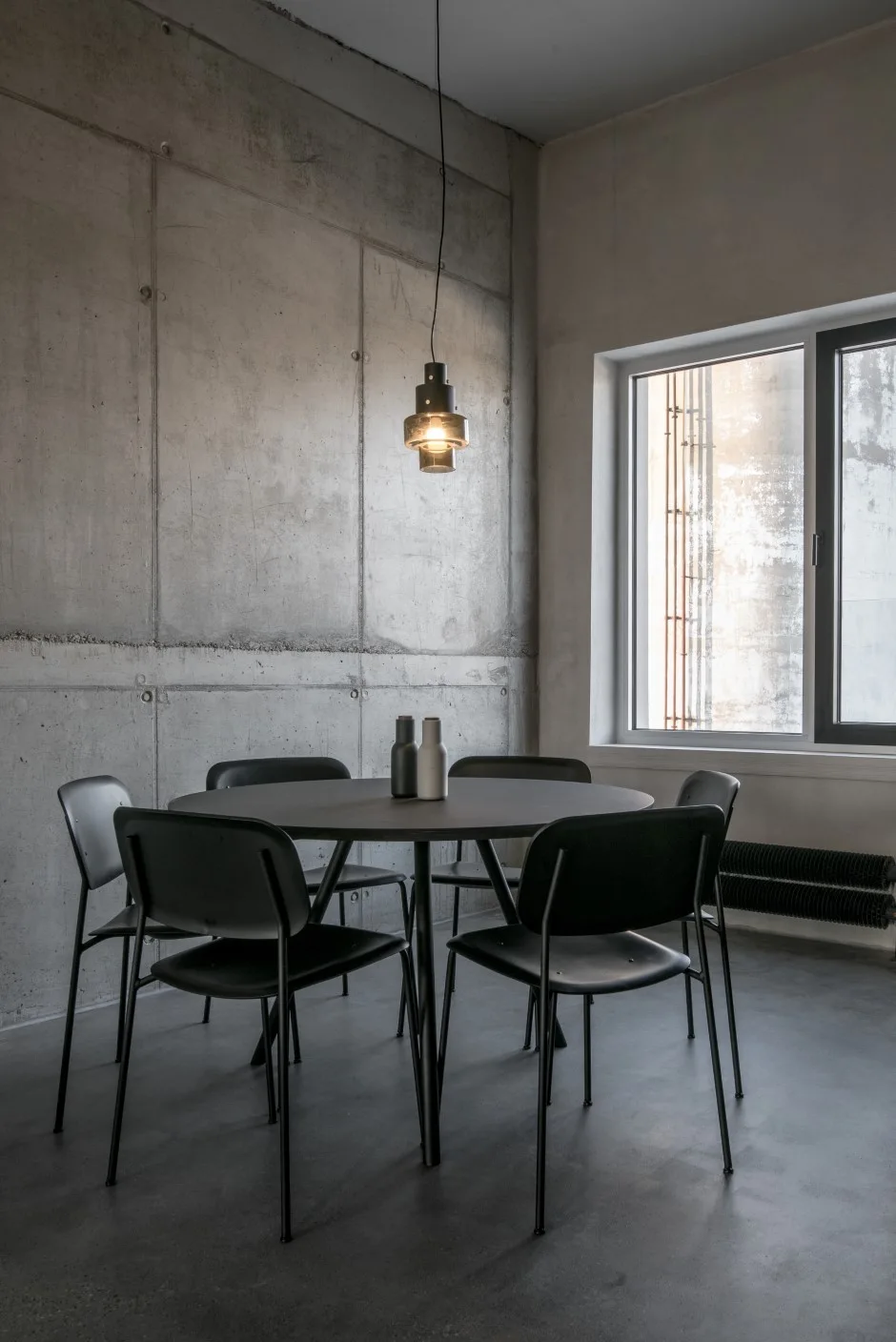 A minimalist dining room features a round black table and chairs set against textured gray concrete walls under a small glass pendant light.
