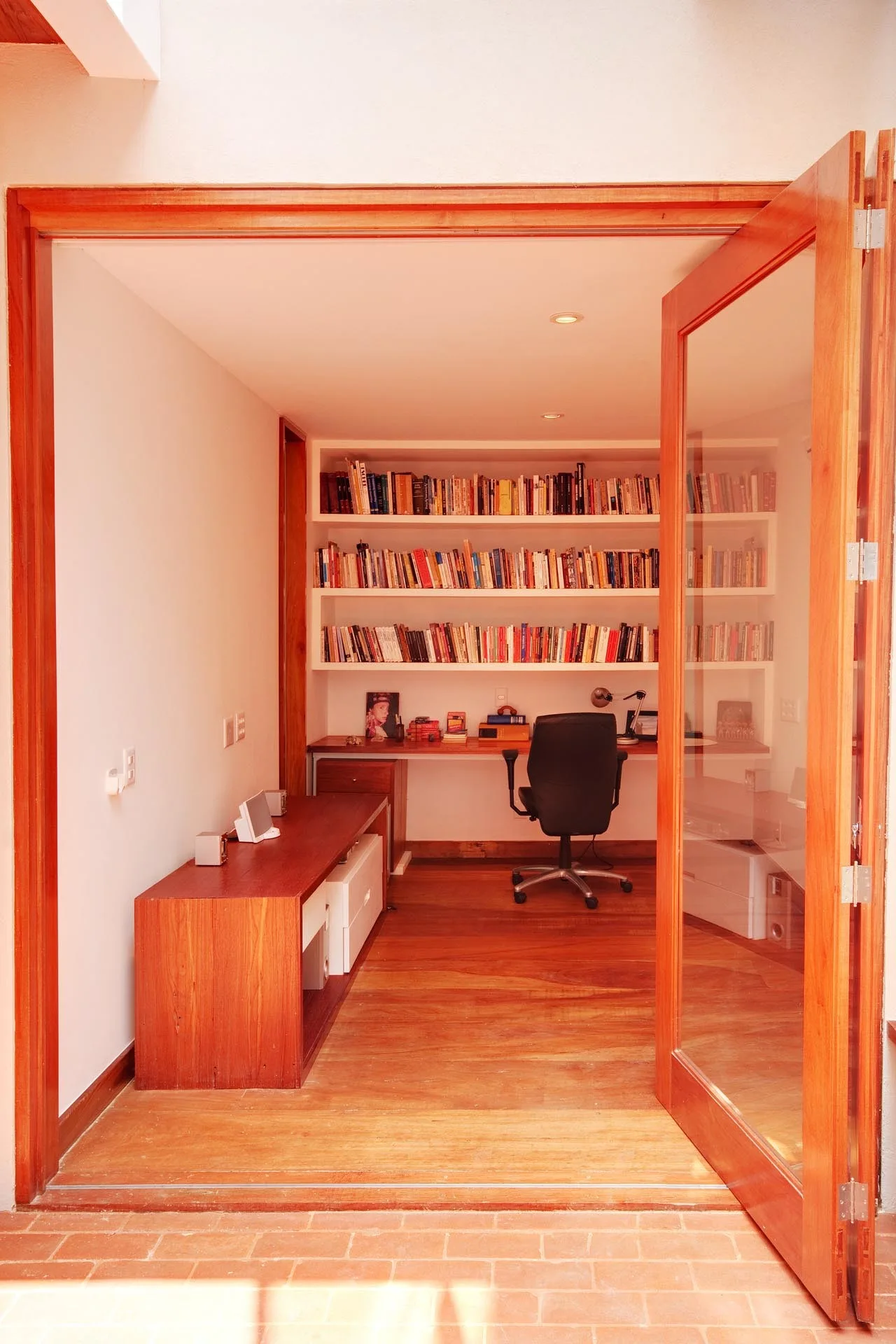 A view through an open wood-framed glass door reveals a home office featuring built-in white bookshelves, a wooden desk, and hardwood floors.