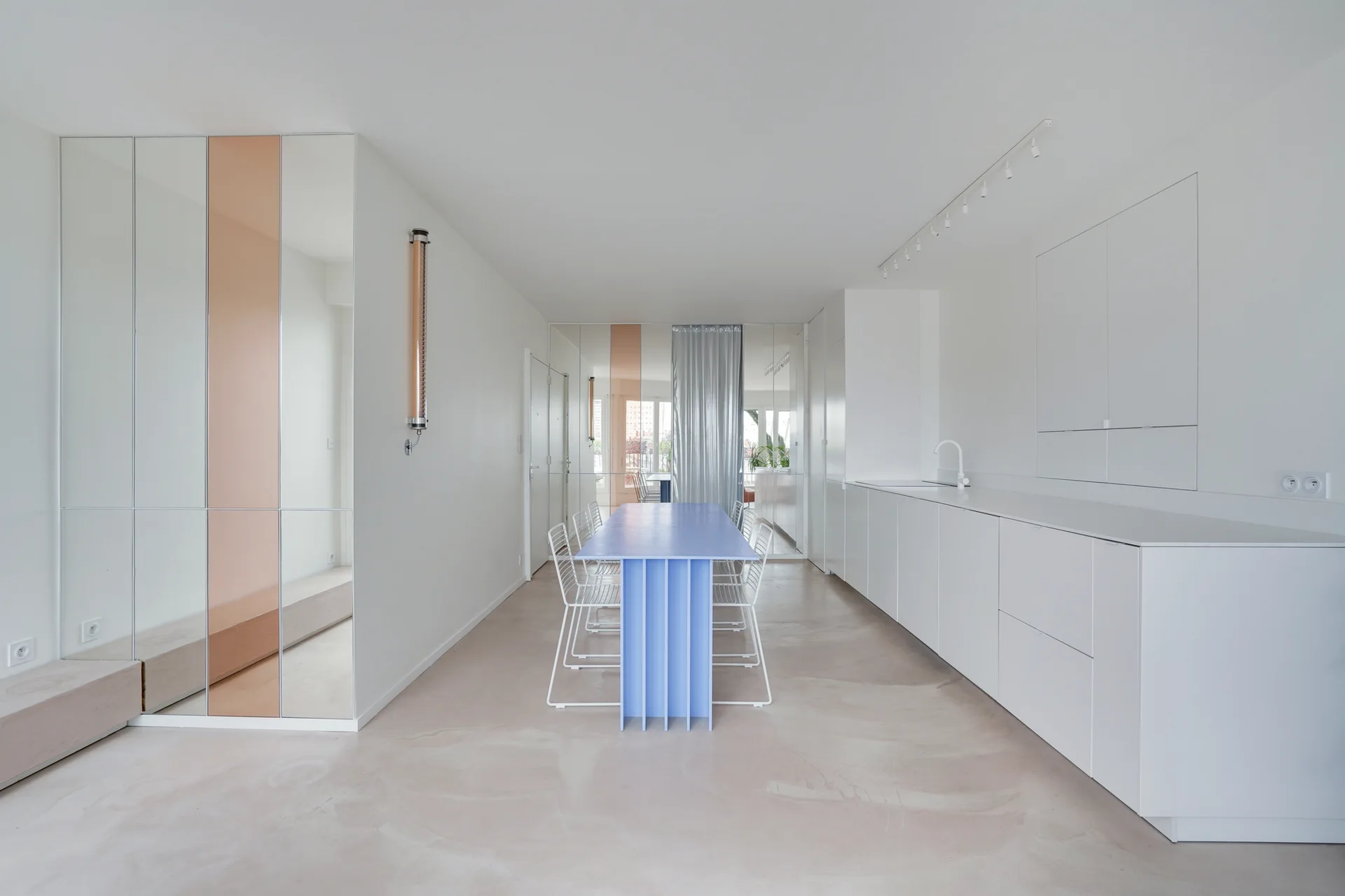 A minimalist dining room features a light blue table surrounded by white wire chairs, adjacent to integrated white cabinetry and mirrored wall panels.