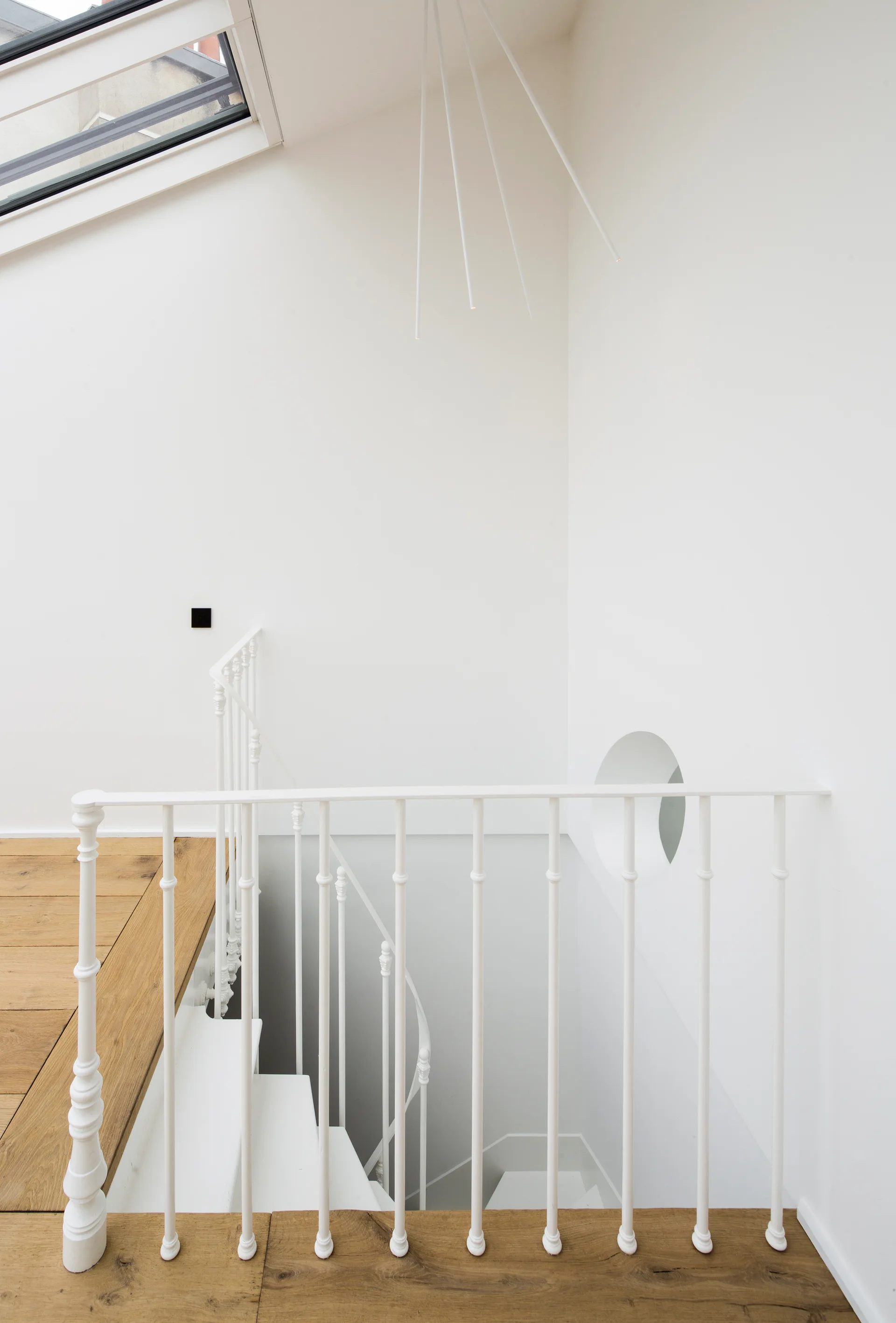 A minimalist white staircase with a metal railing and a round wall cutout features wood plank flooring and a skylight above.