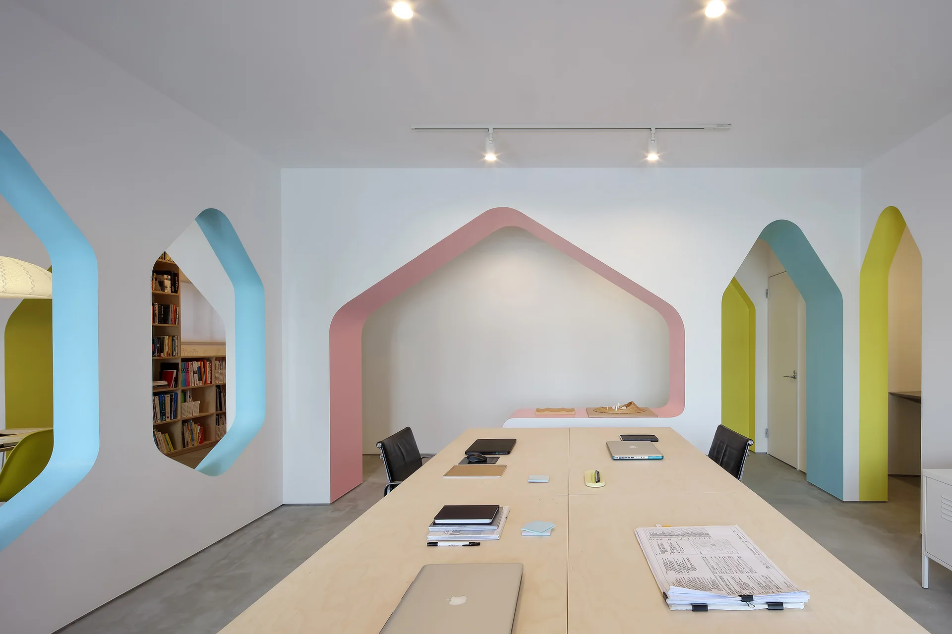 A long light wood conference table stands in a minimalist office featuring white walls with colorful pastel pink, blue, and yellow architectural archways.