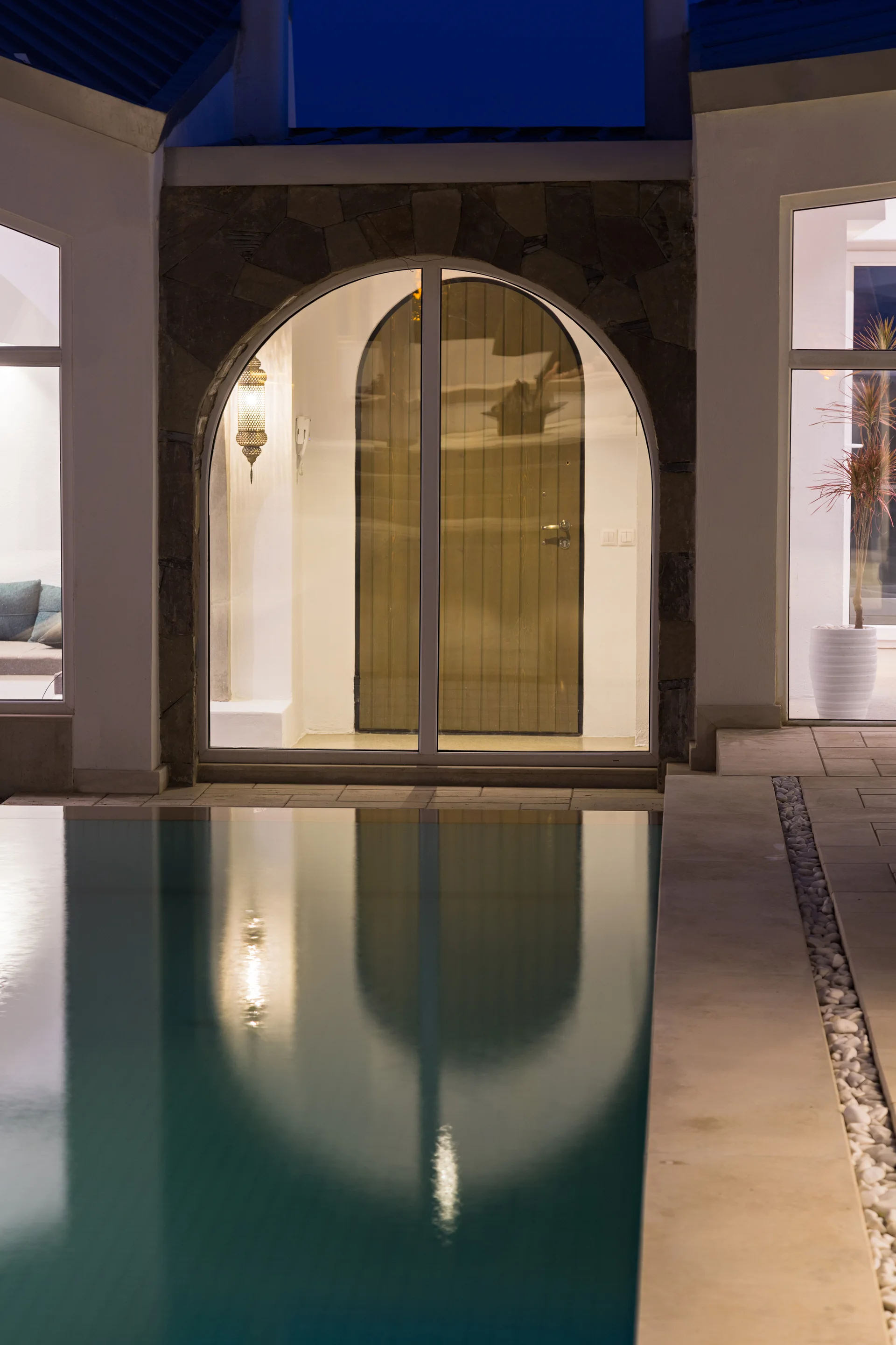 A stone-clad arched entryway with a wooden door overlooks a calm swimming pool reflecting warm ambient light at night.