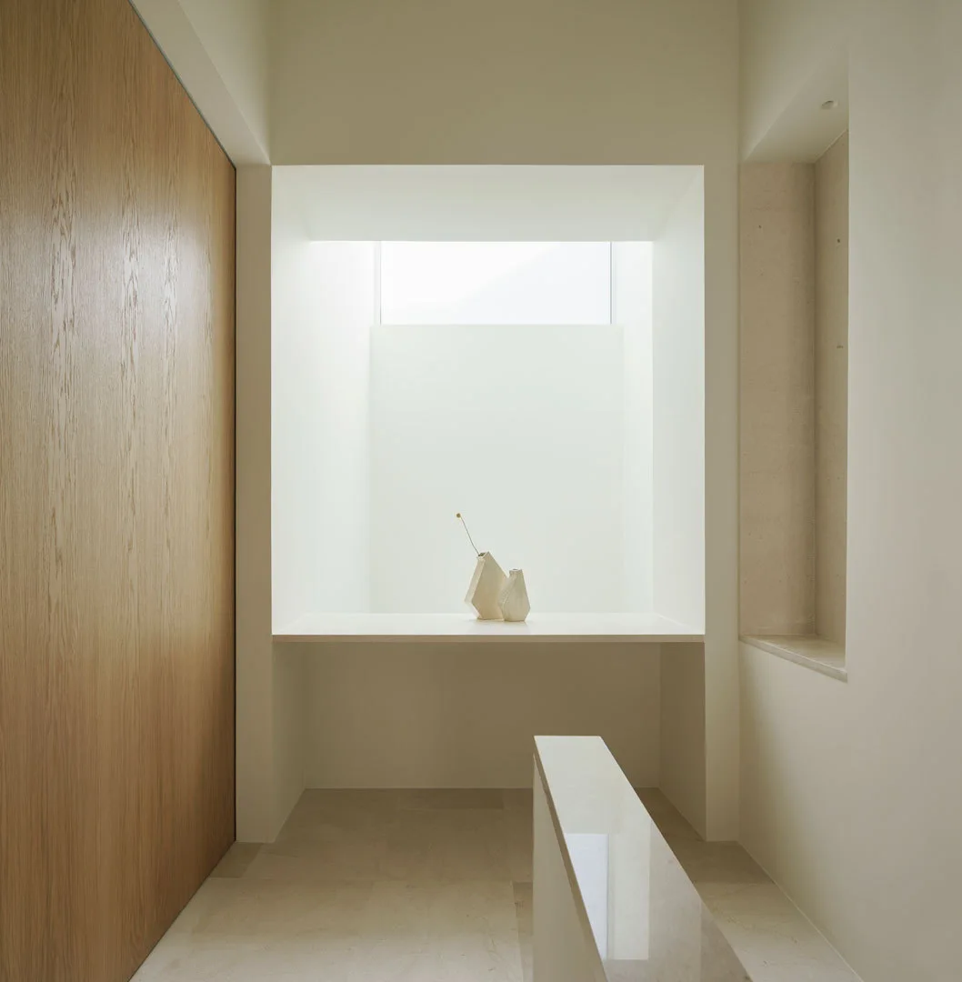 A minimalist corridor with a large wood panel wall, a bright skylight niche displaying small ceramic vases, and beige stone flooring.