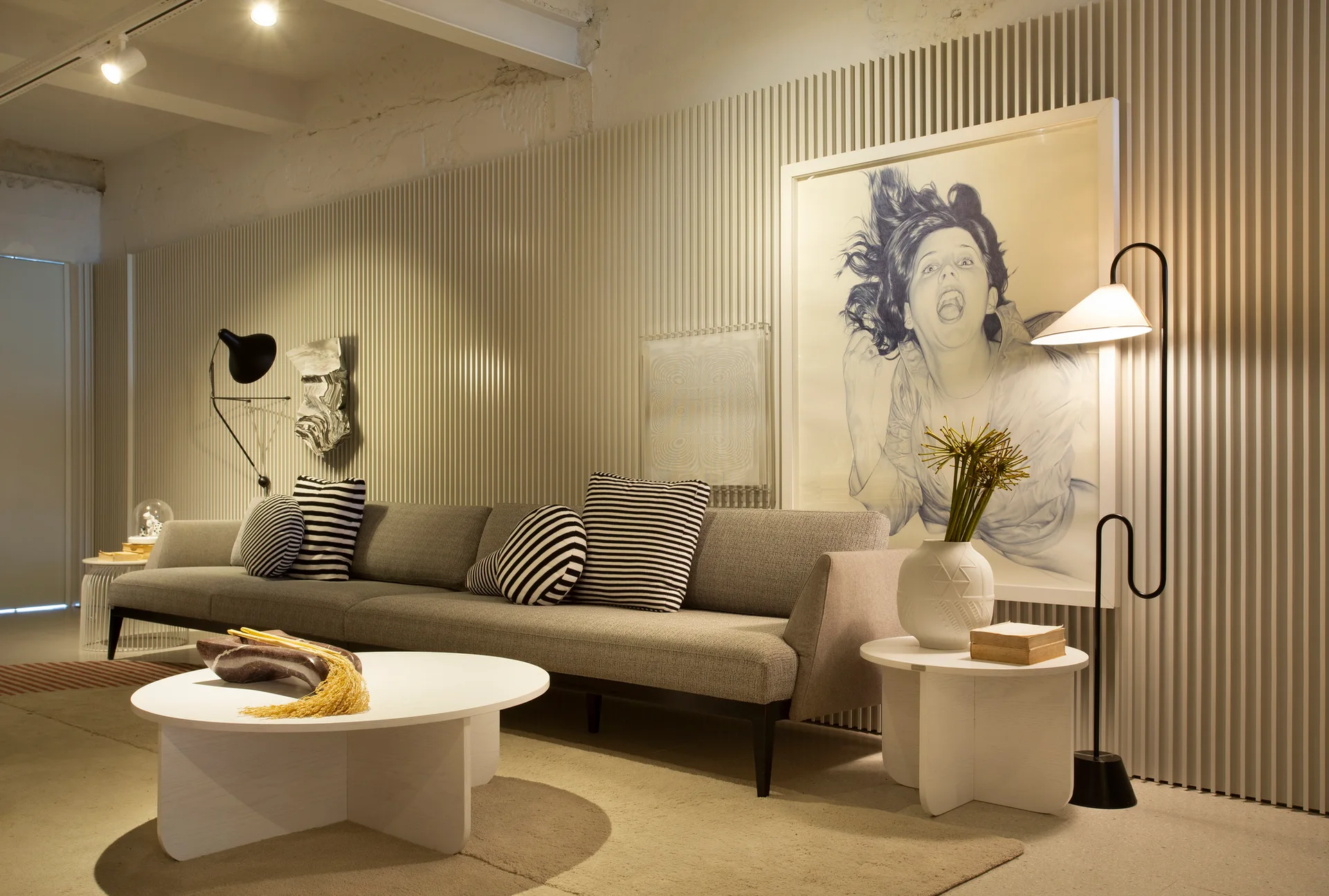 A gray upholstered sofa sits against a cream fluted accent wall alongside round white tables, striped throw pillows, and a large framed portrait.
