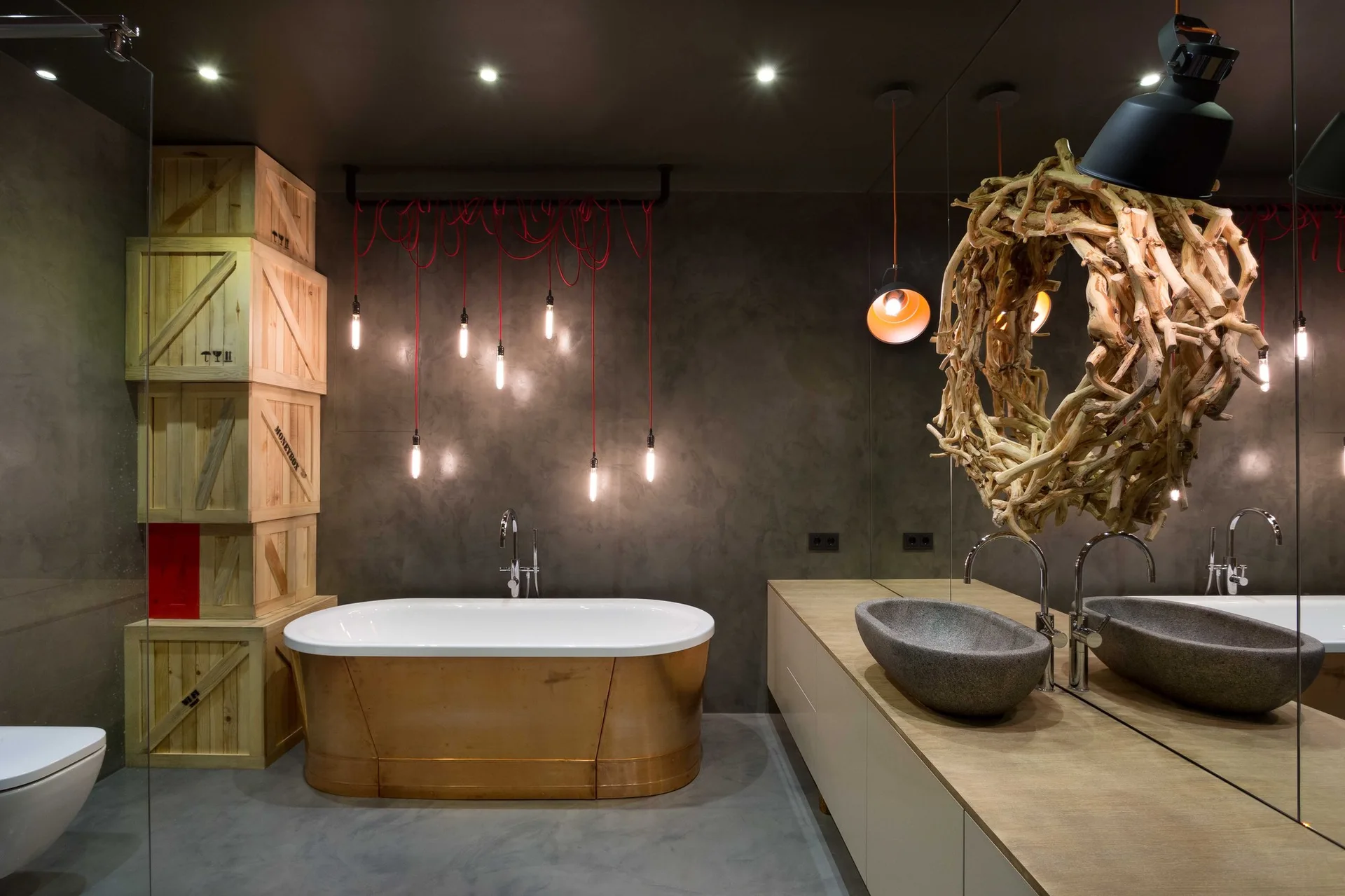 A moody grey bathroom featuring a copper bathtub, stacked wooden crates, hanging red pendant light cords, a stone sink, and a branch-framed mirror.