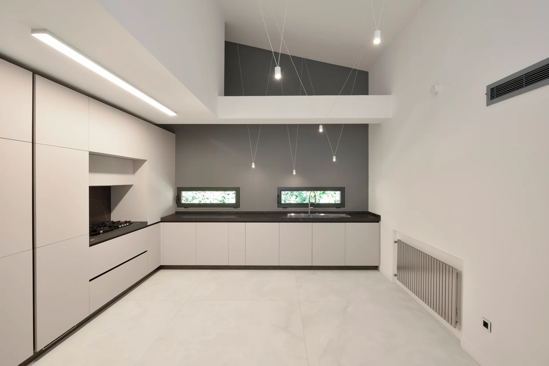 A modern kitchen featuring matte grey cabinets, dark countertops below horizontal windows, and slim pendant lights suspended from a high ceiling.