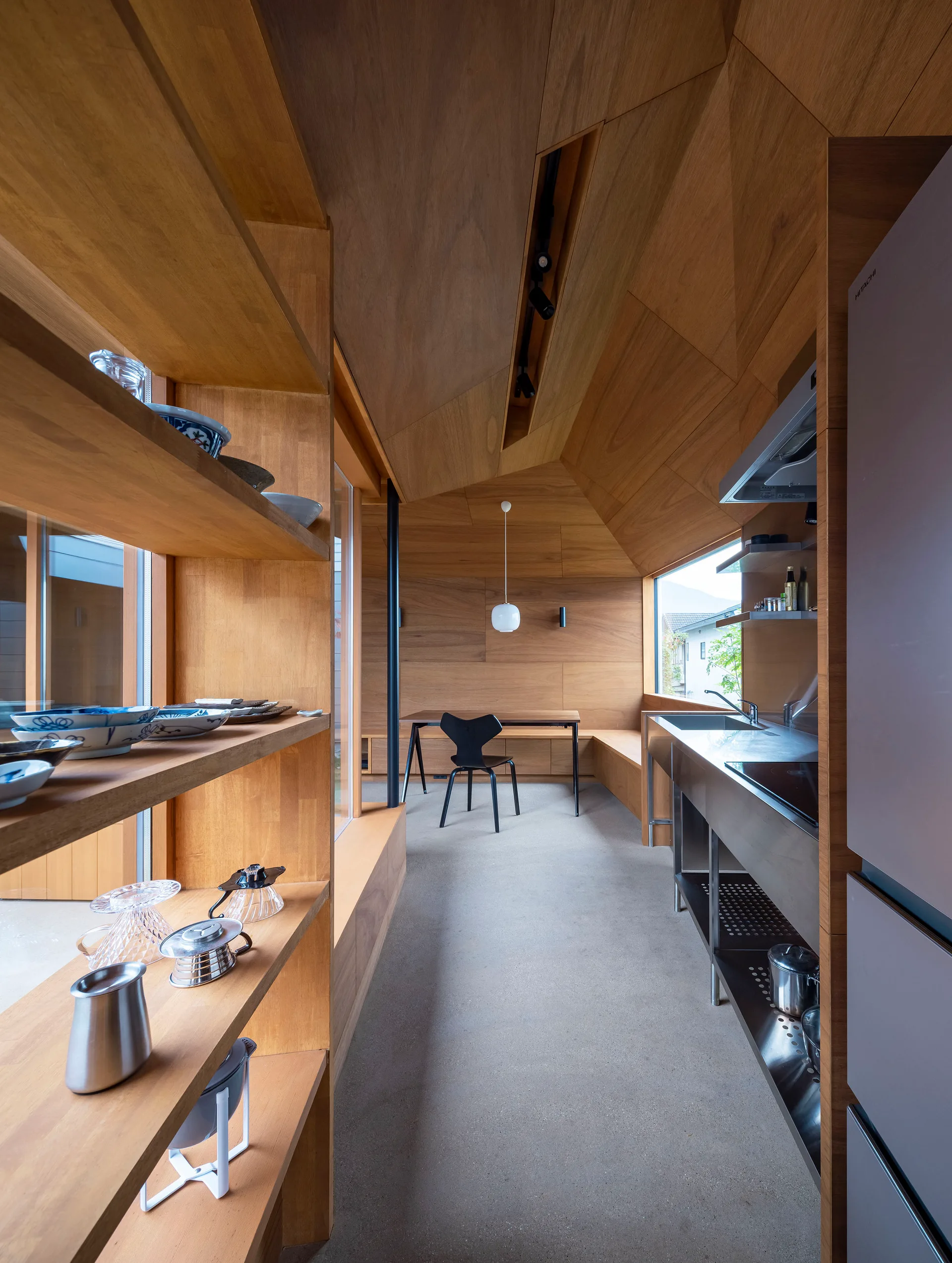 A minimalist wood-paneled kitchen features built-in wooden shelves on the left, a stainless steel counter on the right, and a dining table in the background.