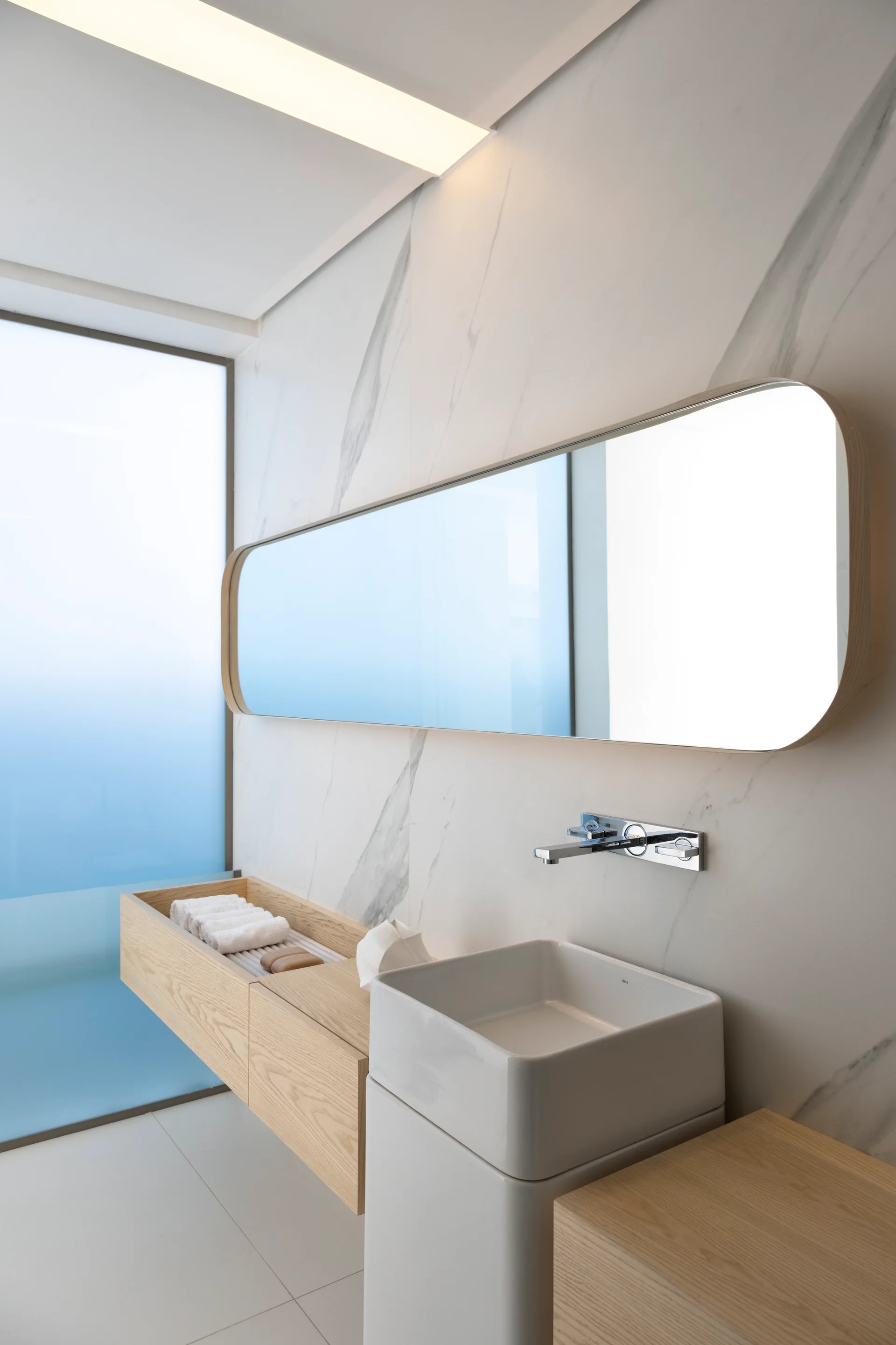 A modern minimalist bathroom featuring a square vessel sink, floating wooden vanity, large oblong mirror, and marble-veined wall tiles.