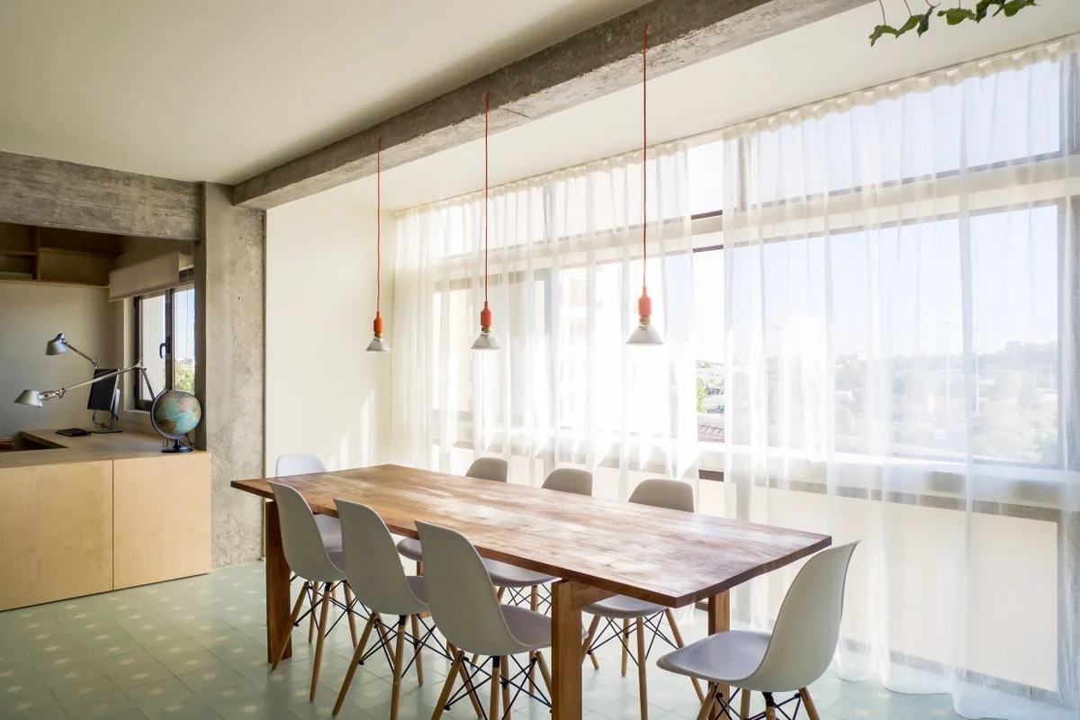A bright dining room features a large wood table, white chairs, hanging pendant lights, and a concrete ceiling beam by a window with sheer curtains.