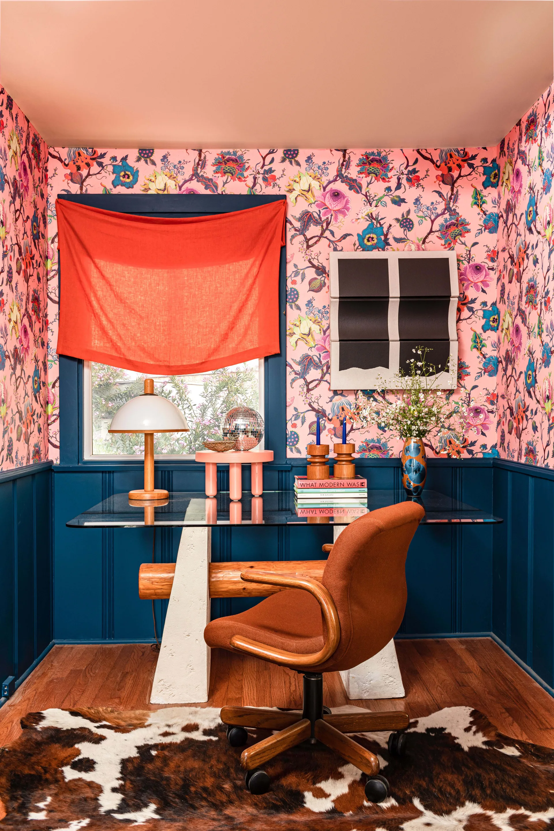 A maximalist home office featuring a glass-top desk, an orange office chair, bold pink floral wallpaper, and dark blue wainscoting.
