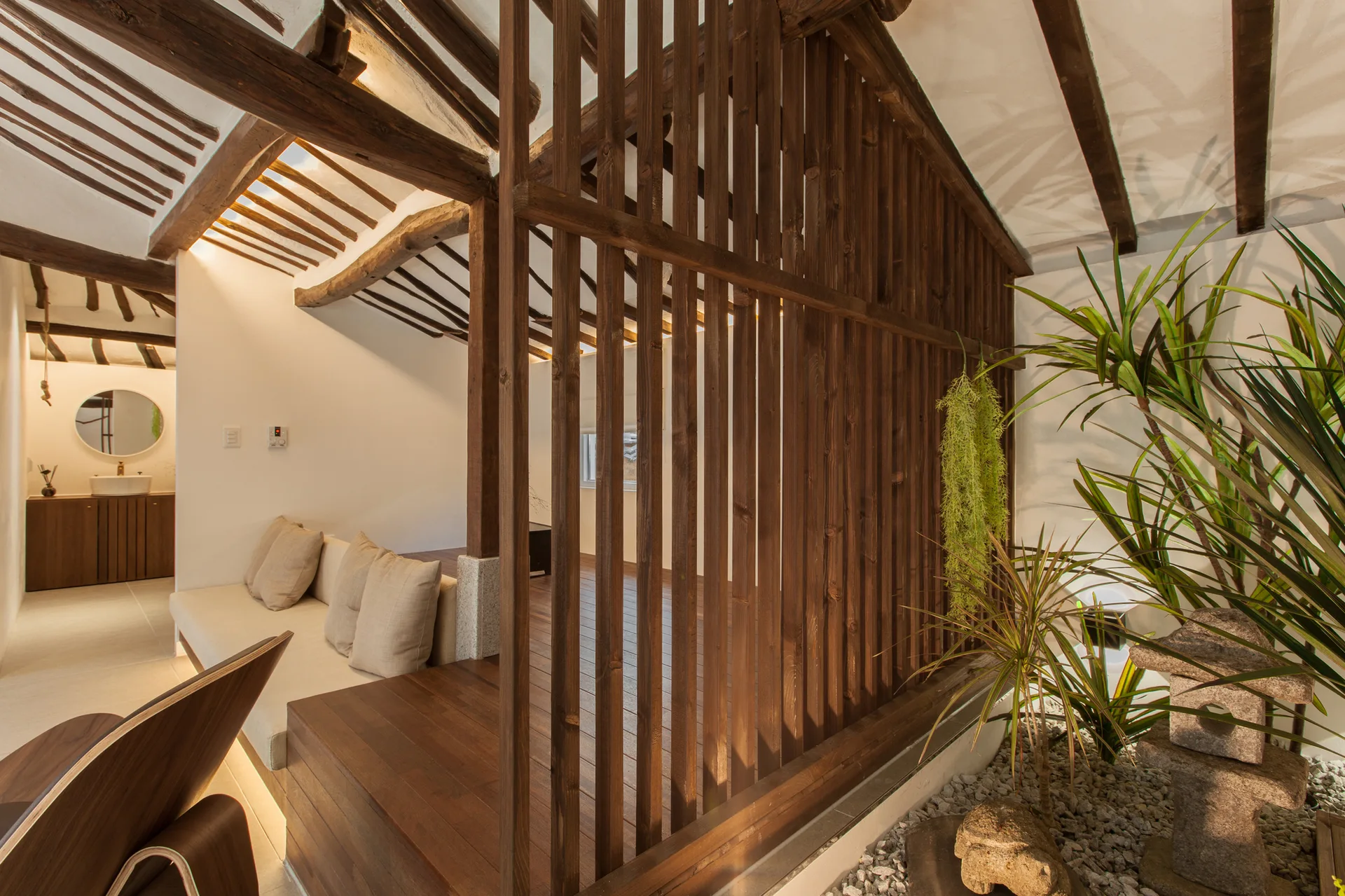 A minimalist living space featuring a dark wooden slat room divider, exposed ceiling beams, a beige sofa, and an indoor pebble garden with plants.