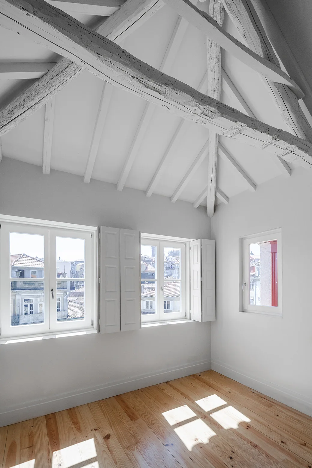 An empty attic room features a white gabled ceiling with exposed timber beams, light wood floors, and white window shutters overlooking city rooftops.