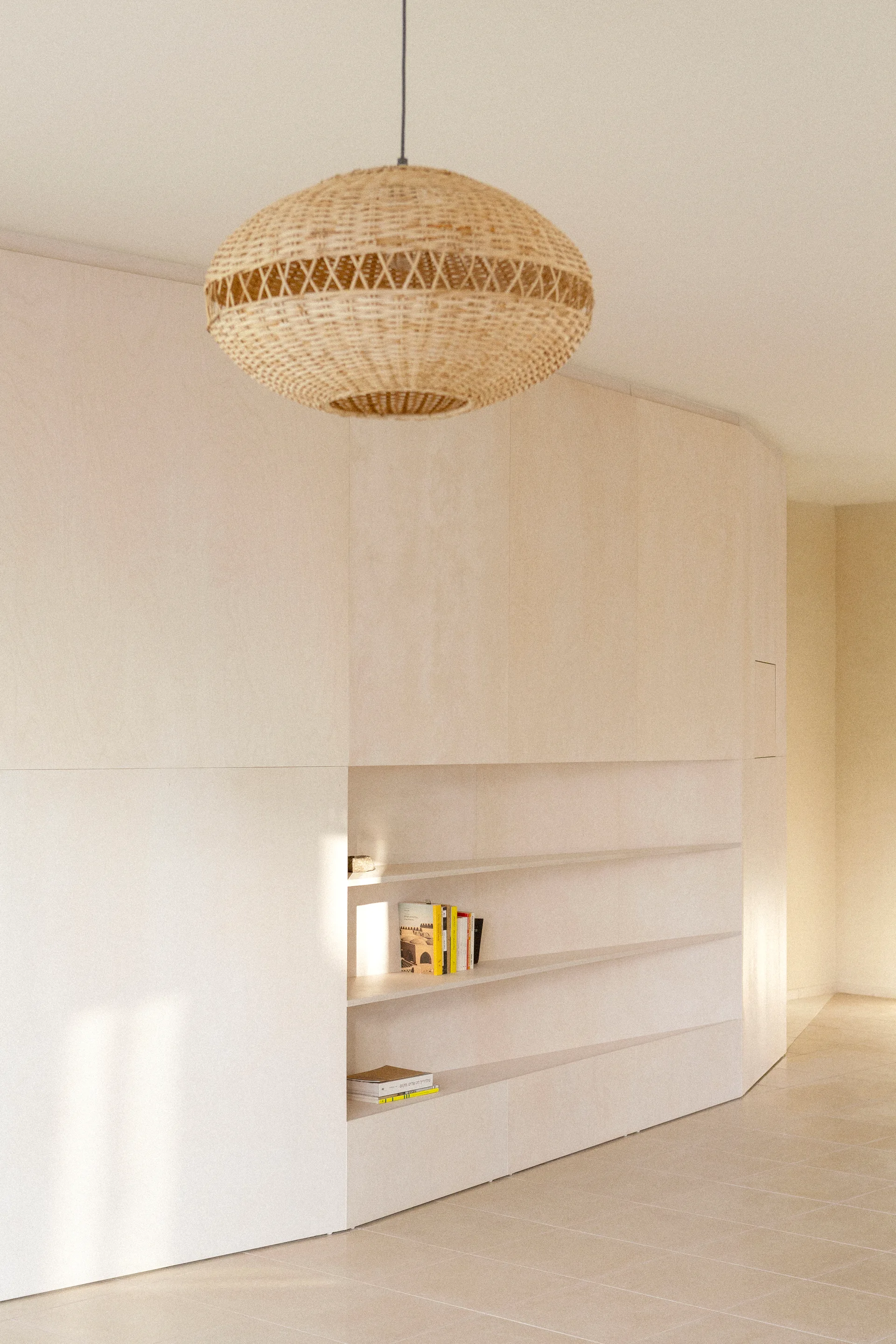 A minimalist light wood storage wall with recessed shelving under a large woven wicker pendant light.