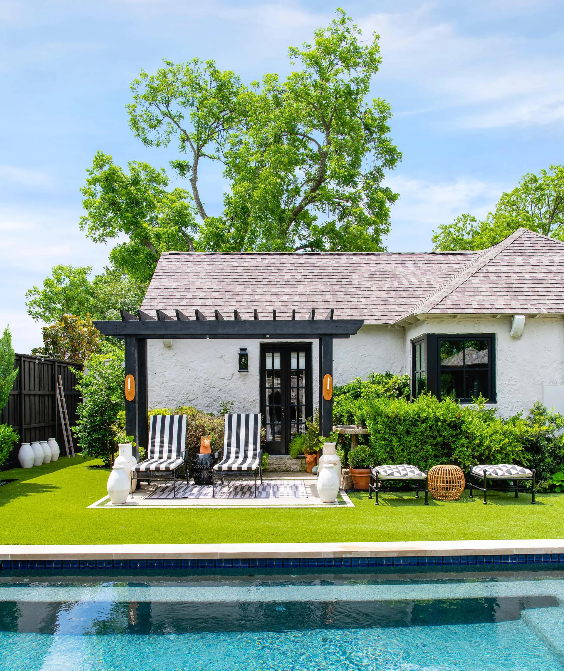 Black-and-white striped lounge chairs sit under a dark wood pergola on a lawn next to a swimming pool and a white stucco house.