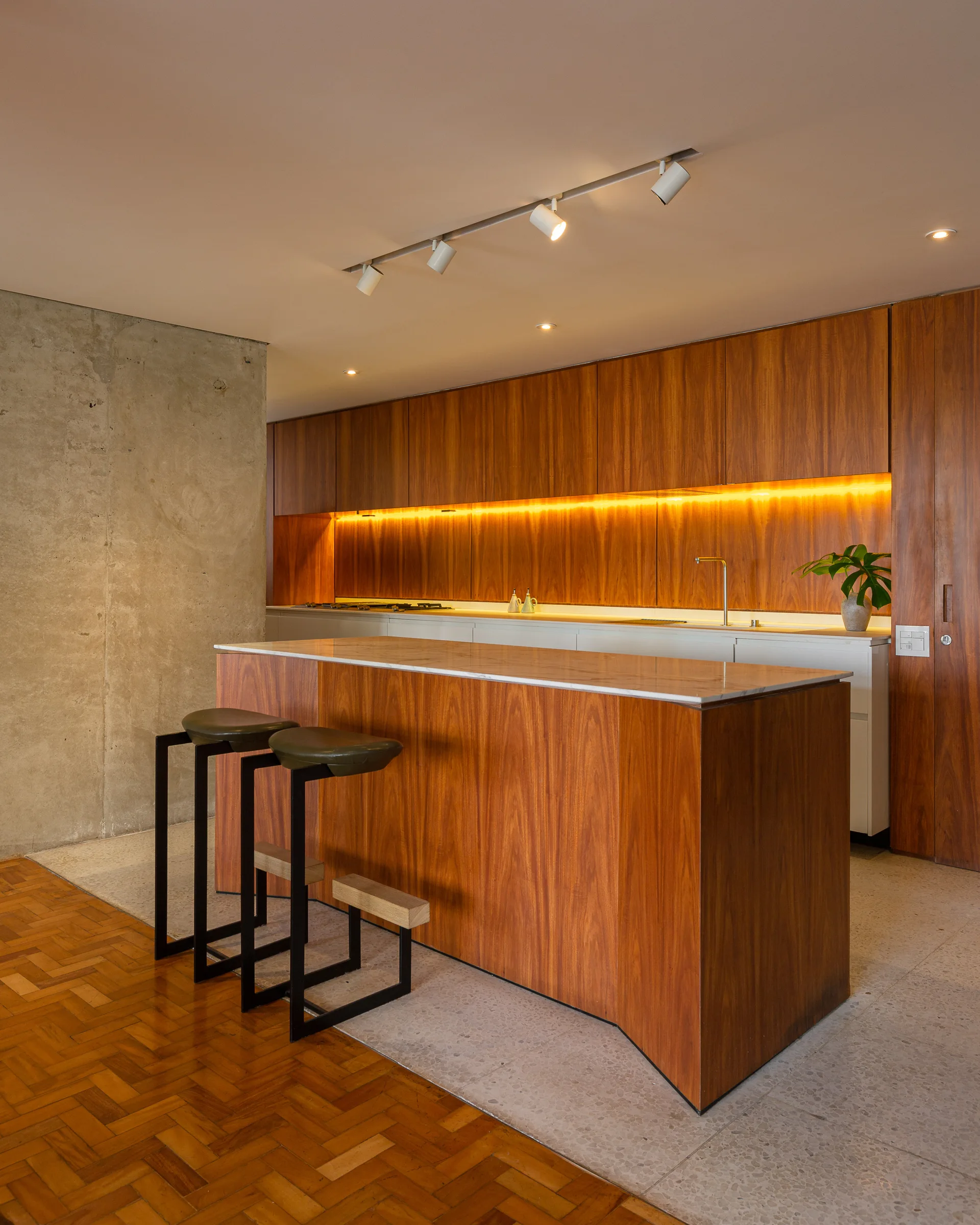 A modern kitchen featuring rich wood cabinetry, a matching island with a stone countertop, black bar stools, and an exposed concrete wall.