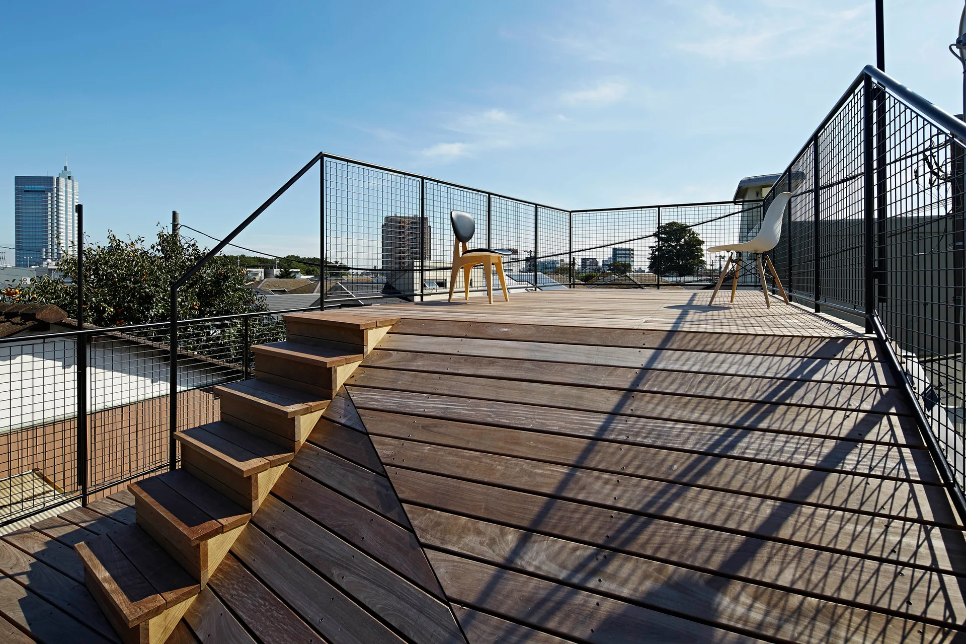 A multi-level wooden rooftop deck features built-in stairs, black wire mesh railing, and two minimalist chairs overlooking a city.