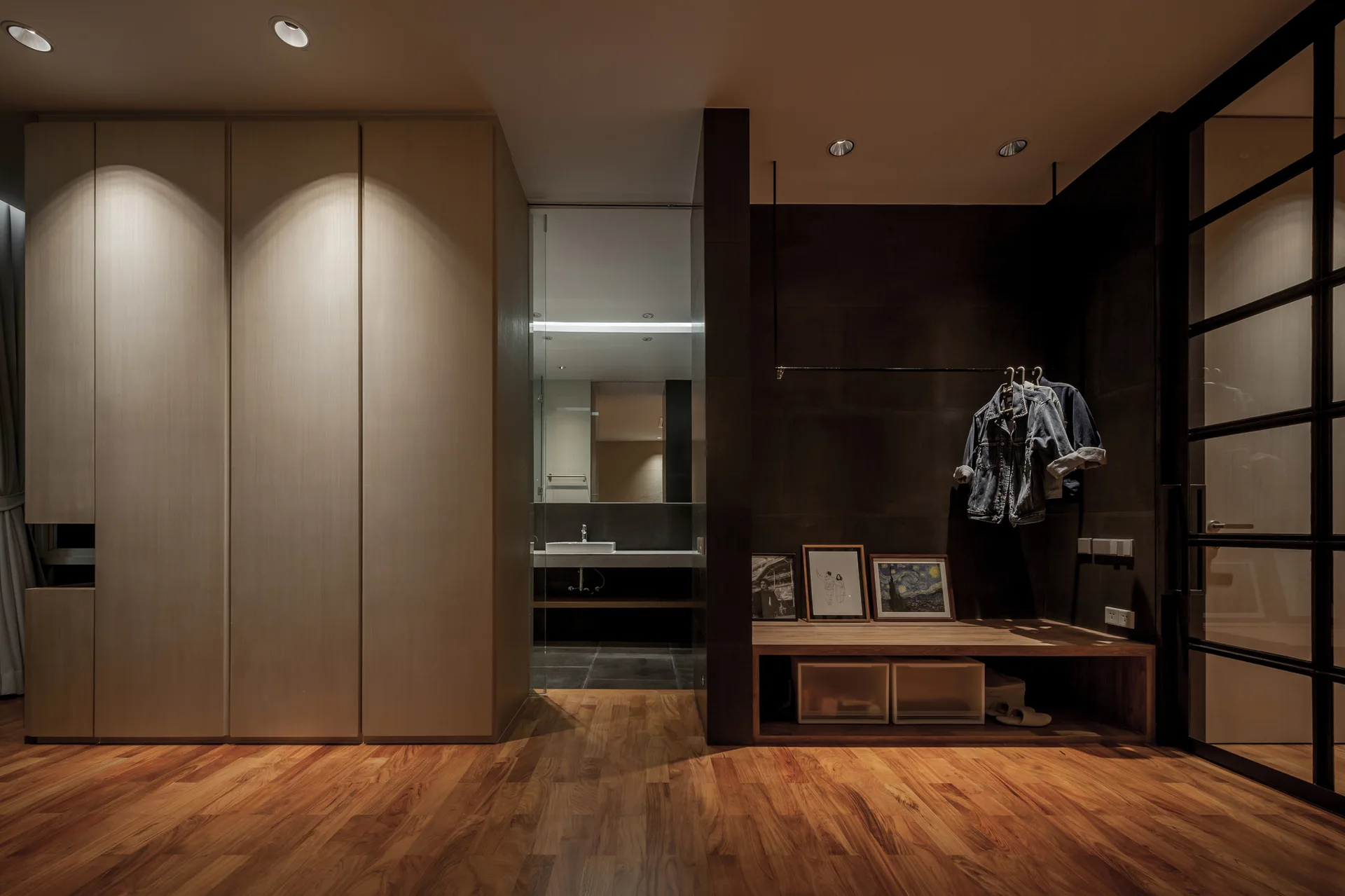 A minimalist dressing area featuring light wood wardrobes, a dark accent wall with a wood bench and hanging rack, and a view of a bathroom.