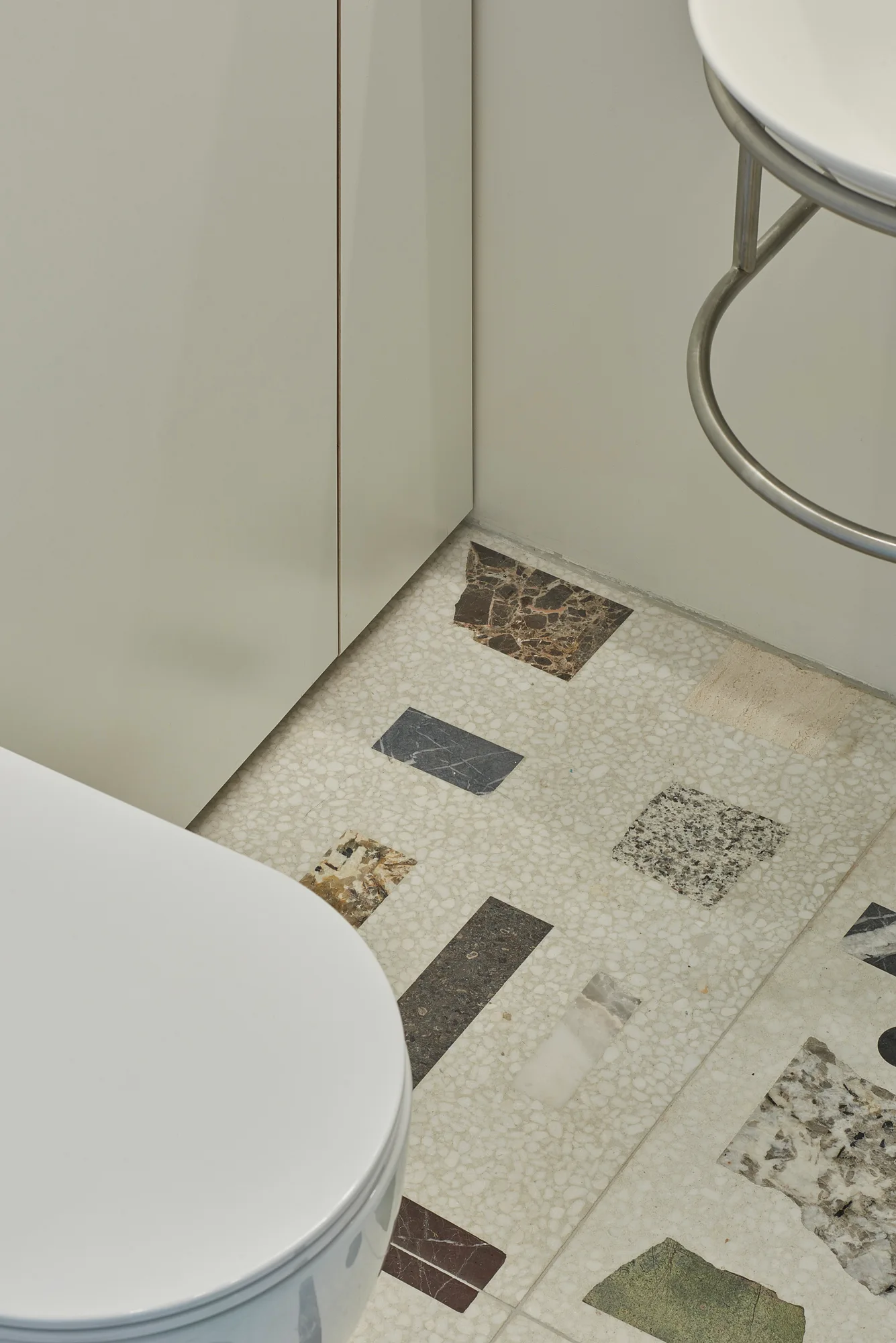 A close-up of a bathroom floor featuring custom terrazzo tiles with inlaid marble fragments next to a white toilet and minimalist beige cabinet.
