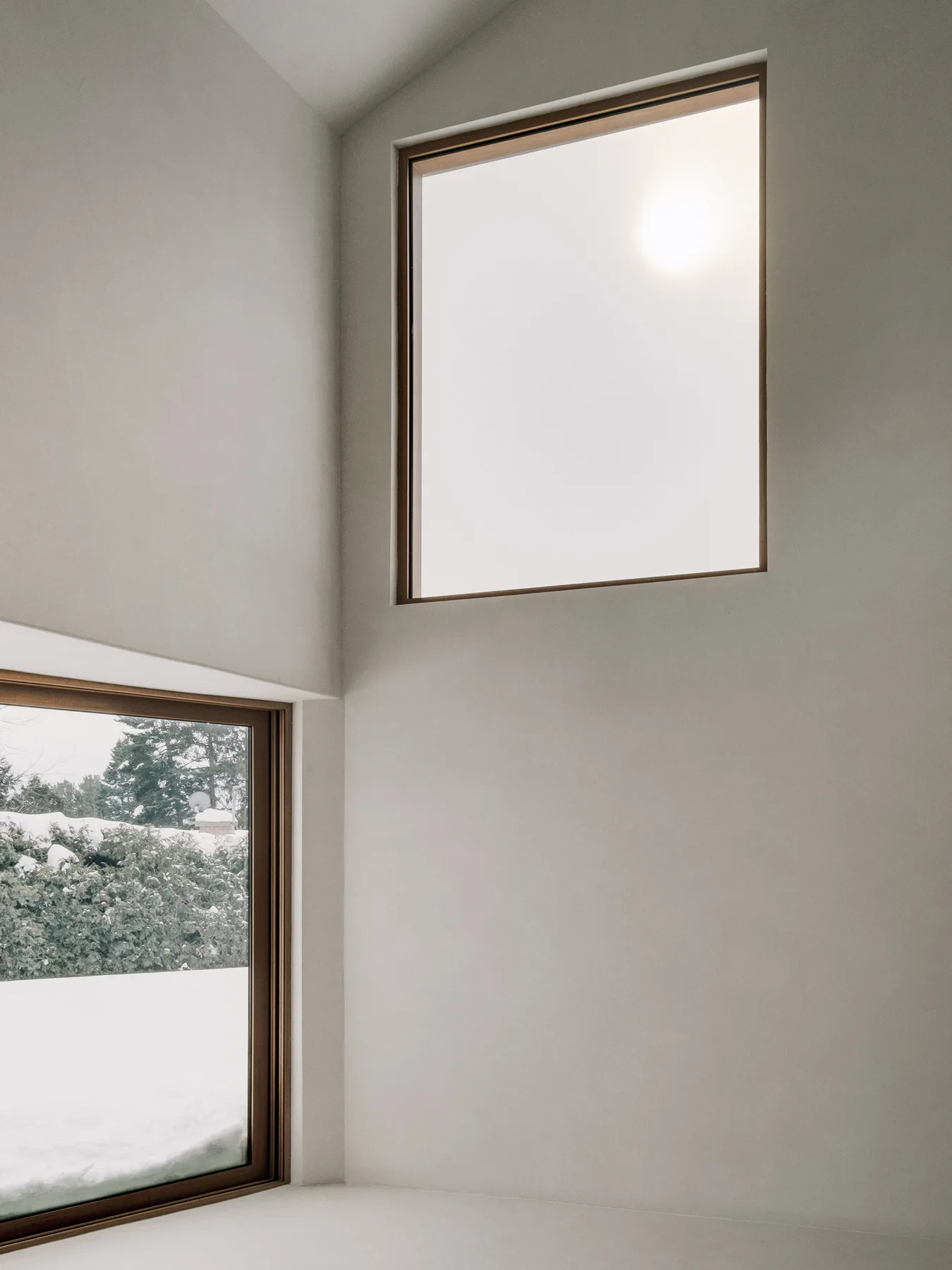 A minimalist interior corner featuring smooth off-white plaster walls, a sloped ceiling, and two wood-framed windows looking out onto snow.